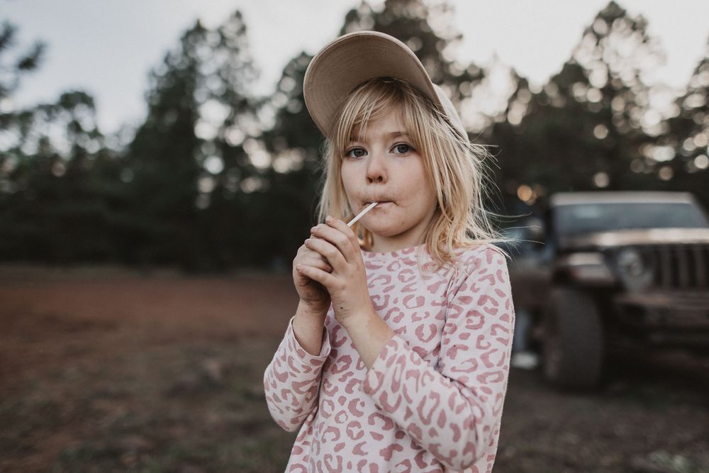 Young girl in pink leopard print sweater and tan cap holds a lollipop outdoors near a Jeep.