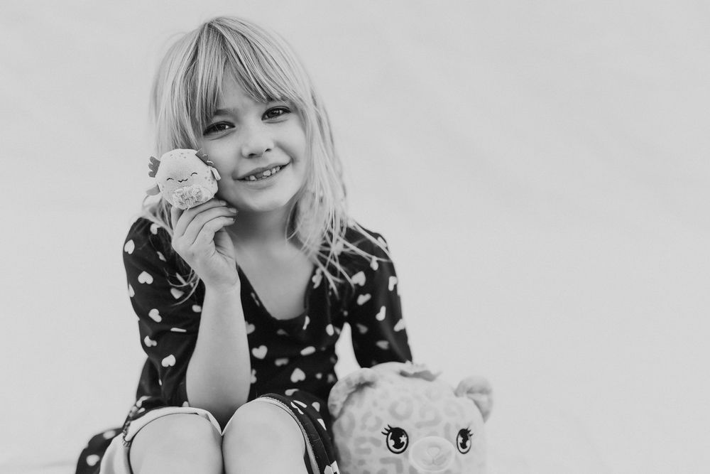 Black and white photo of a smiling young girl holding a small toy while sitting with a stuffed animal cat.