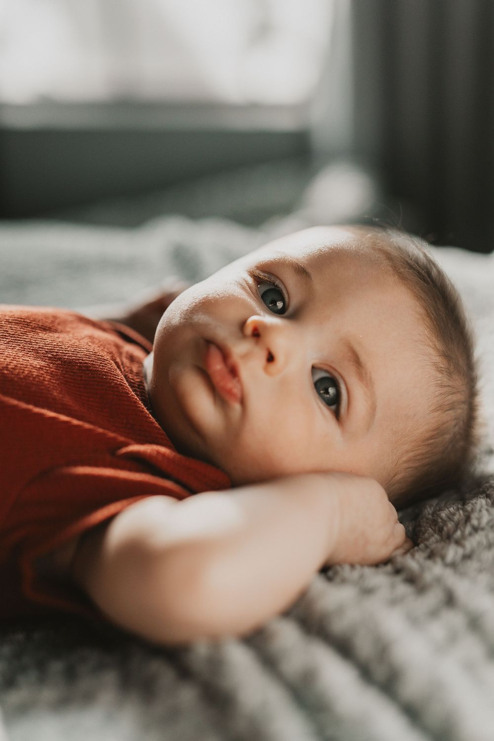 A cute baby in a red onesie lying on a gray blanket, gazing upward with bright eyes in soft natural light.