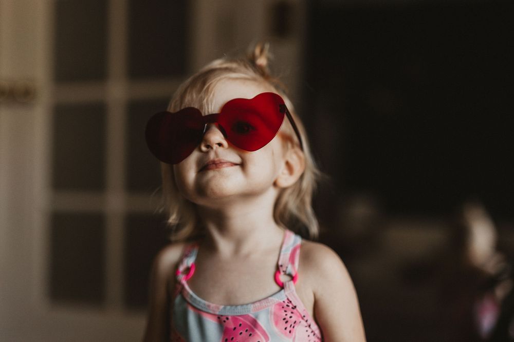 A joyful toddler girl wearing red heart-shaped sunglasses smiles while wearing a pink floral swimsuit indoors.