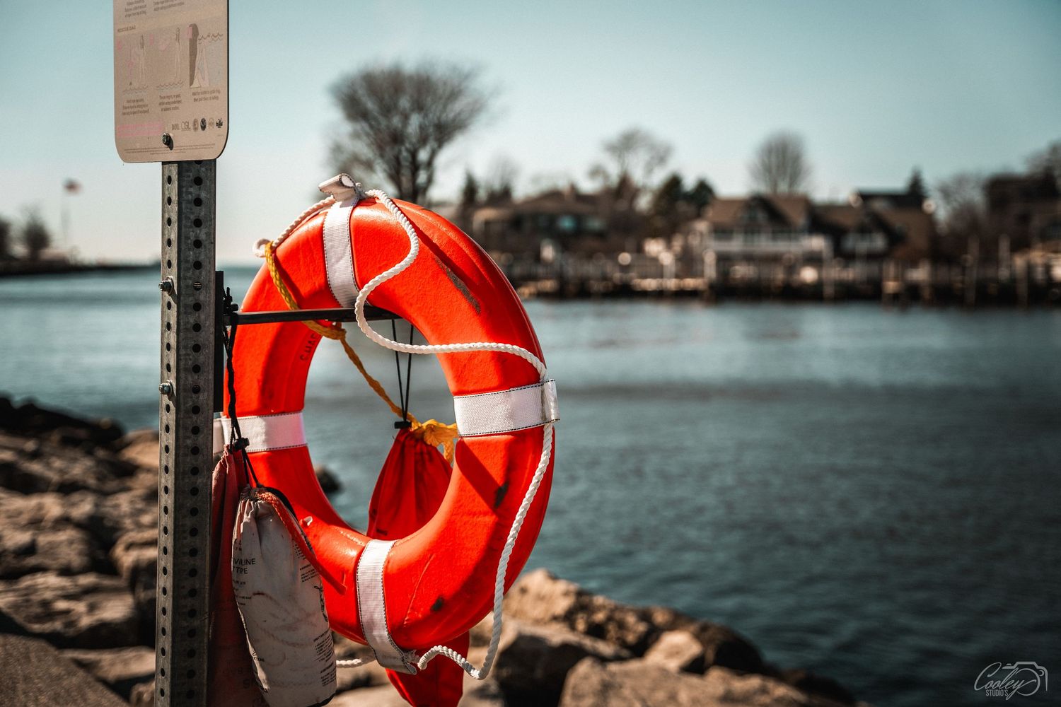 Orange life preserver ring mounted on a post near a rocky waterfront with town in background.