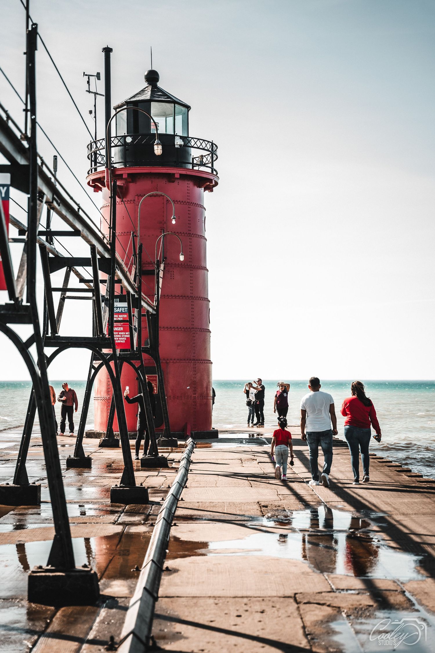 Families walking along a wet pier toward a red lighthouse on a bright sunny day.