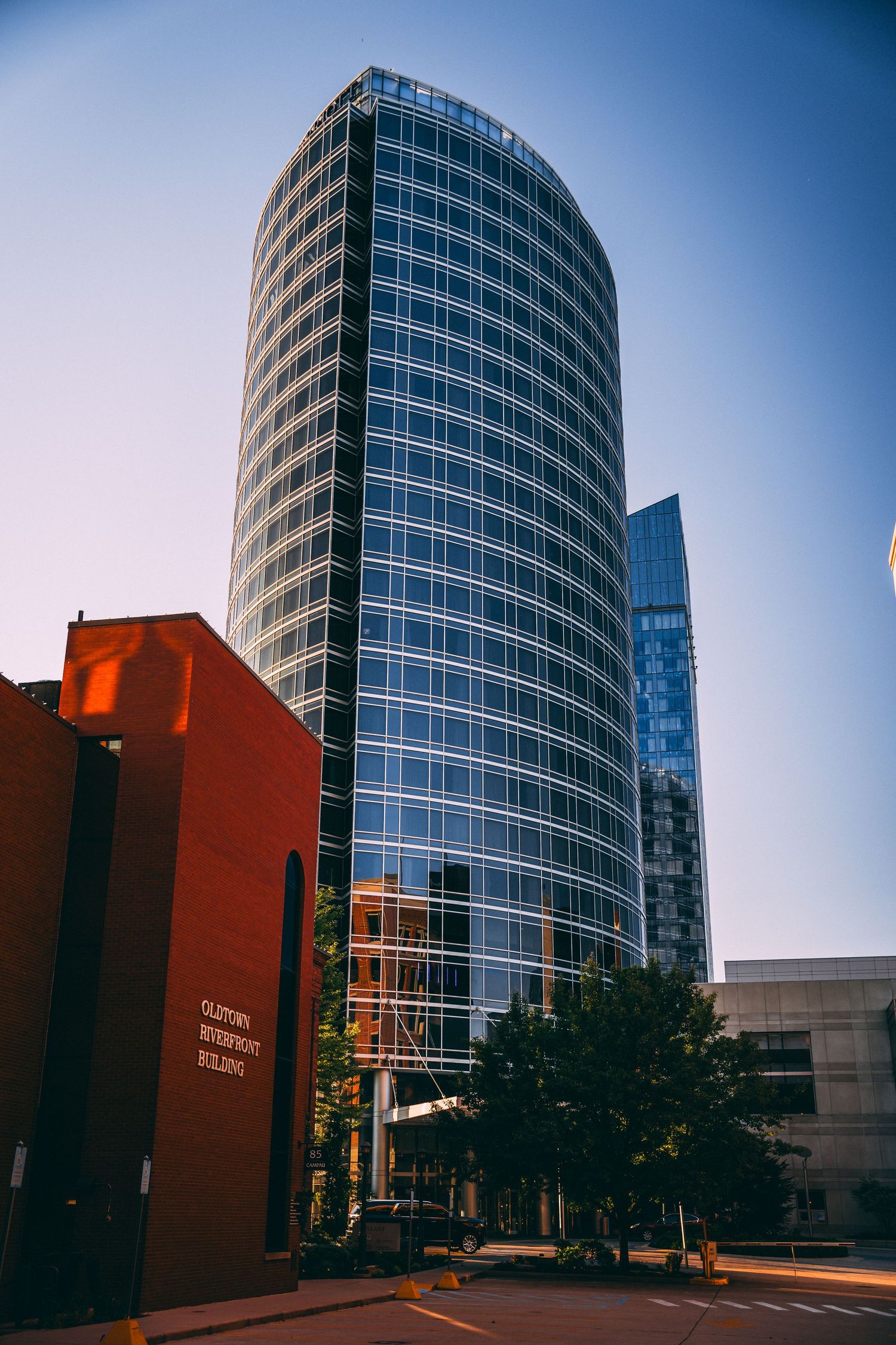 Modern cylindrical glass skyscraper towering over red brick building in downtown city.