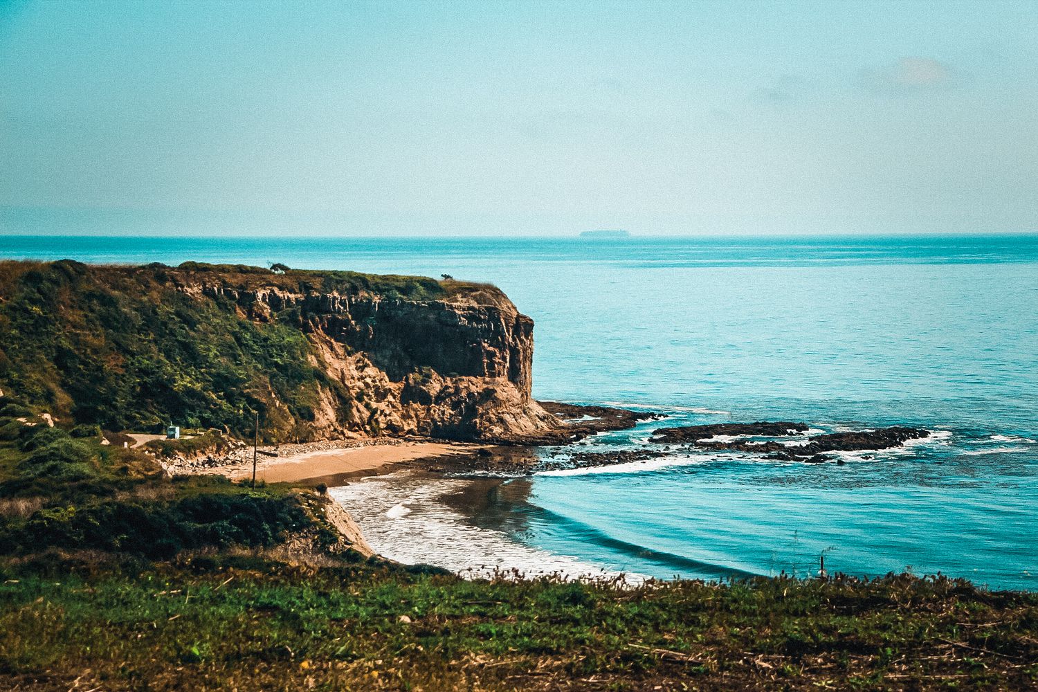 Scenic coastal cliff overlooking a turquoise ocean with a small sandy beach below.
