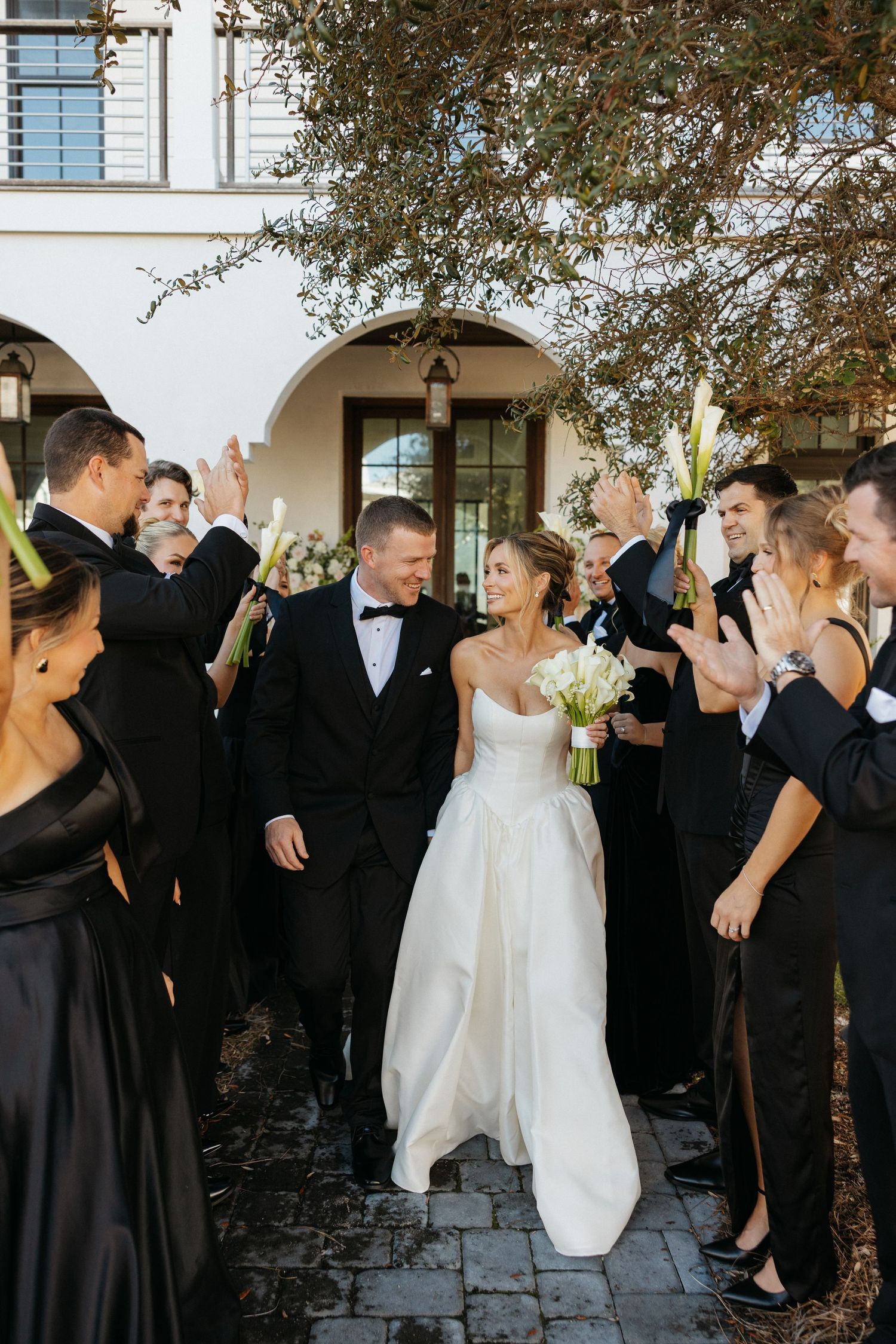 Wedding party in black attire celebrates with couple outside white arched building during daytime ceremony.
