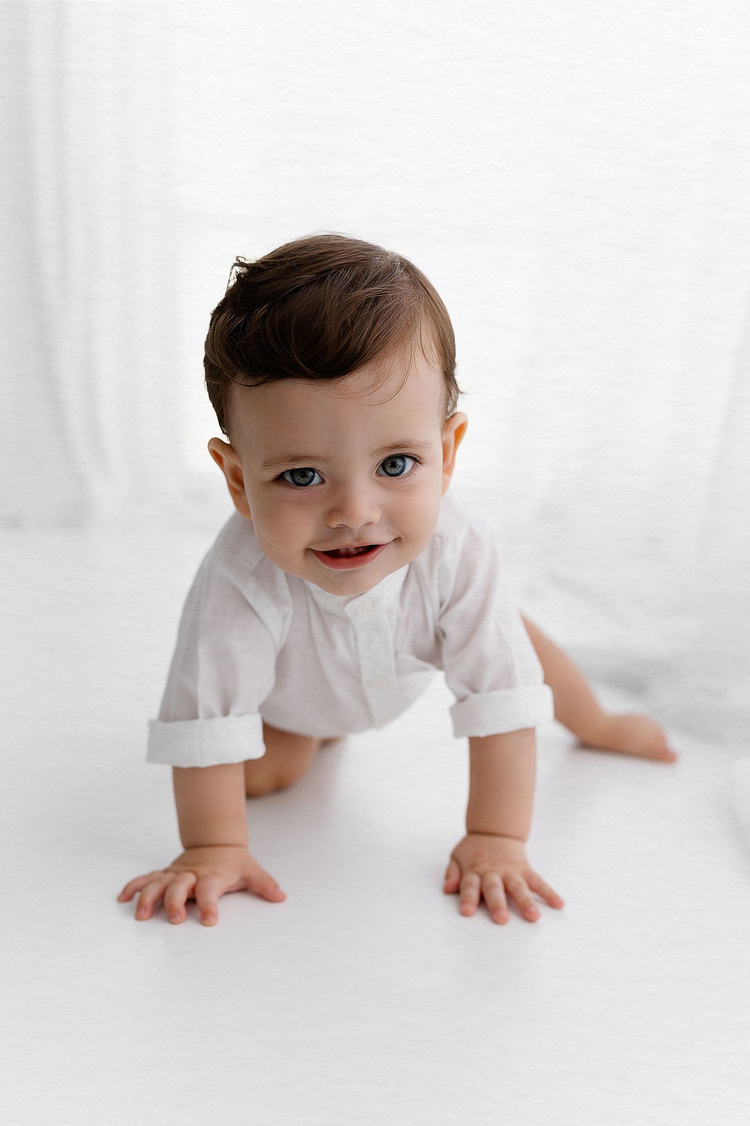 A baby in white clothing crawls on a white background, showing a bright smile.