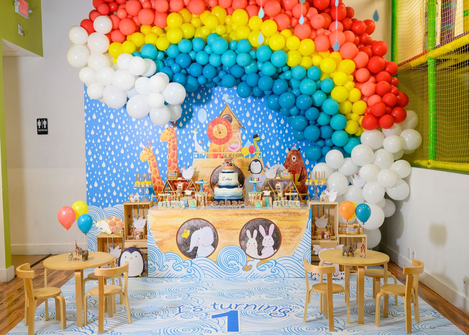 Colorful rainbow balloon arch decorates a party setup with wooden chairs and a dessert table against a blue backdrop.