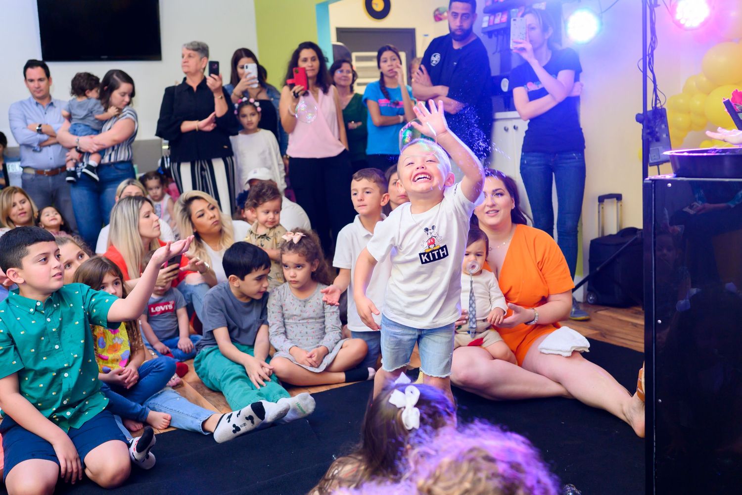 Group of people gathered on floor enjoying entertainment with colorful stage lights and musical equipment visible.