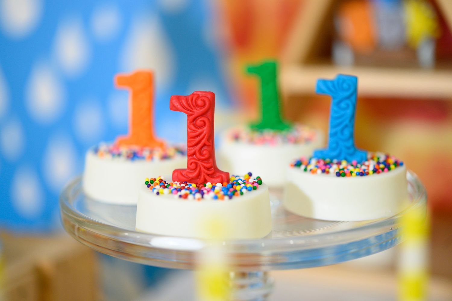 Four colorful number one candles with sprinkles sit atop white frosted cookies against a blue polka dot background.