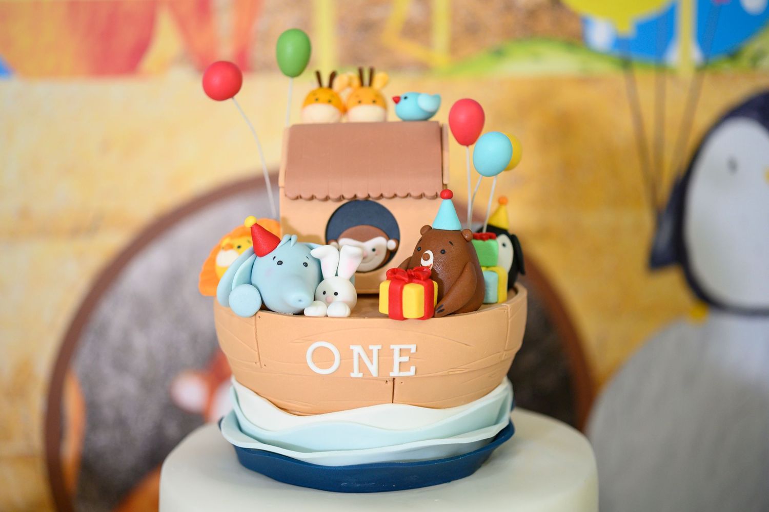 A decorated first birthday cake with balloons and animal decorations on a table against a yellow background.