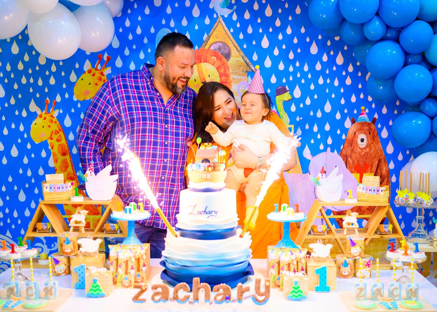A festive birthday party setup with blue balloon decorations, a white tiered cake, and teddy bear decorations on the table.