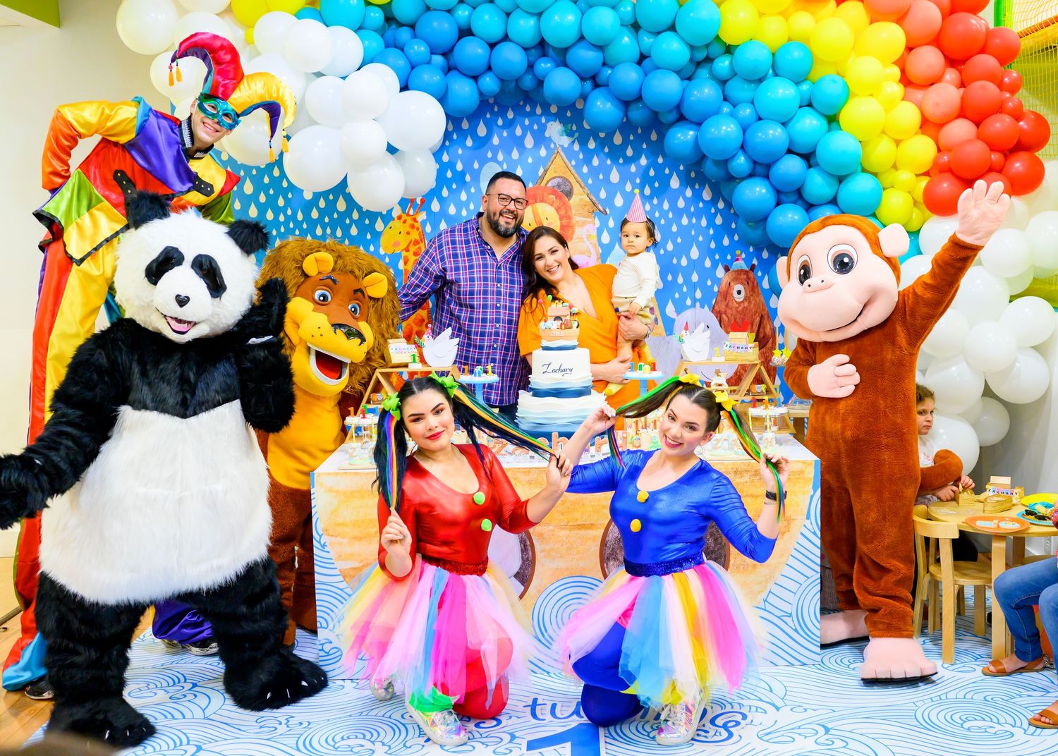 Colorful rainbow balloon arch decorates party scene with mascot characters and dancers in bright tutu costumes.