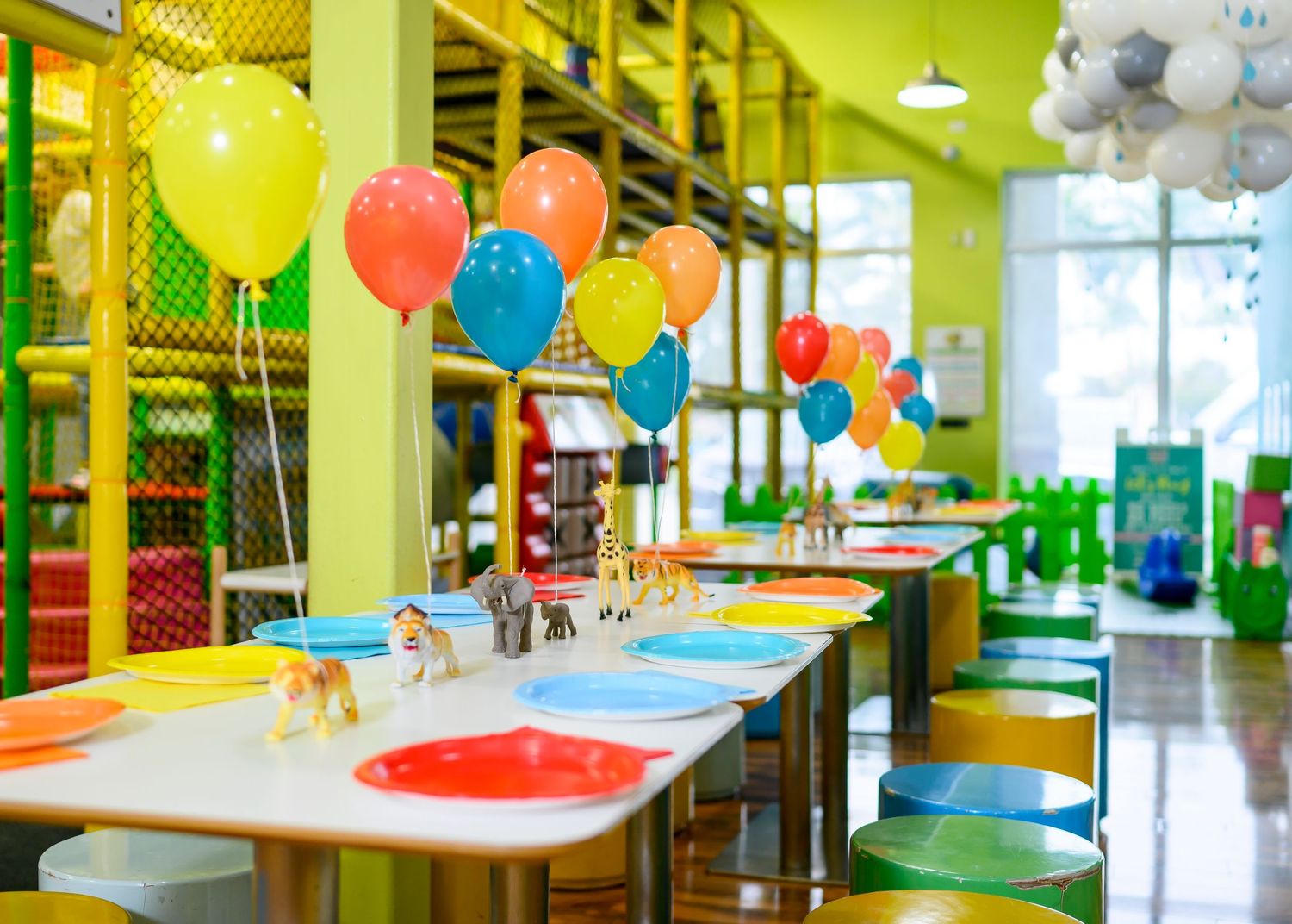 Colorful balloons decorate a children's party room with long tables and green walls in an indoor play space.