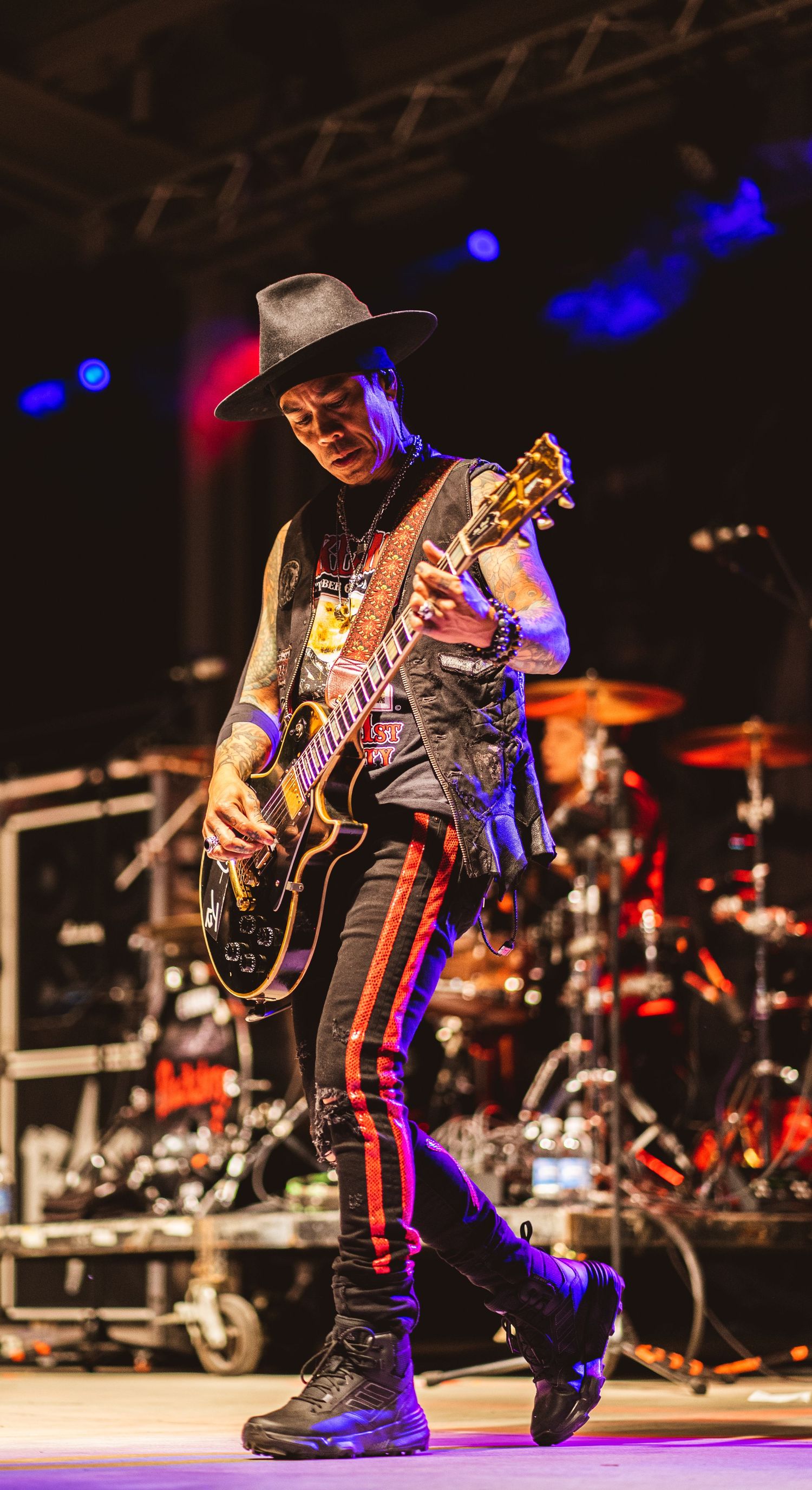 Guitarist in cowboy hat and striped pants performs on stage with dramatic red and blue lighting.