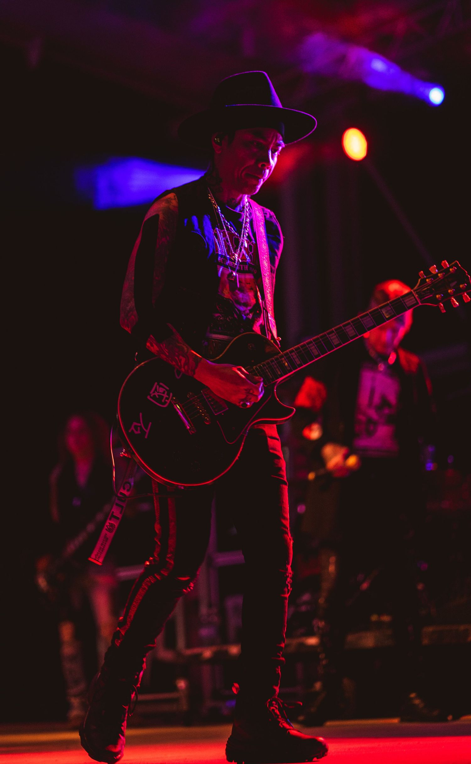 Guitar player in dark clothing performs on dimly lit stage with red and blue spotlights.