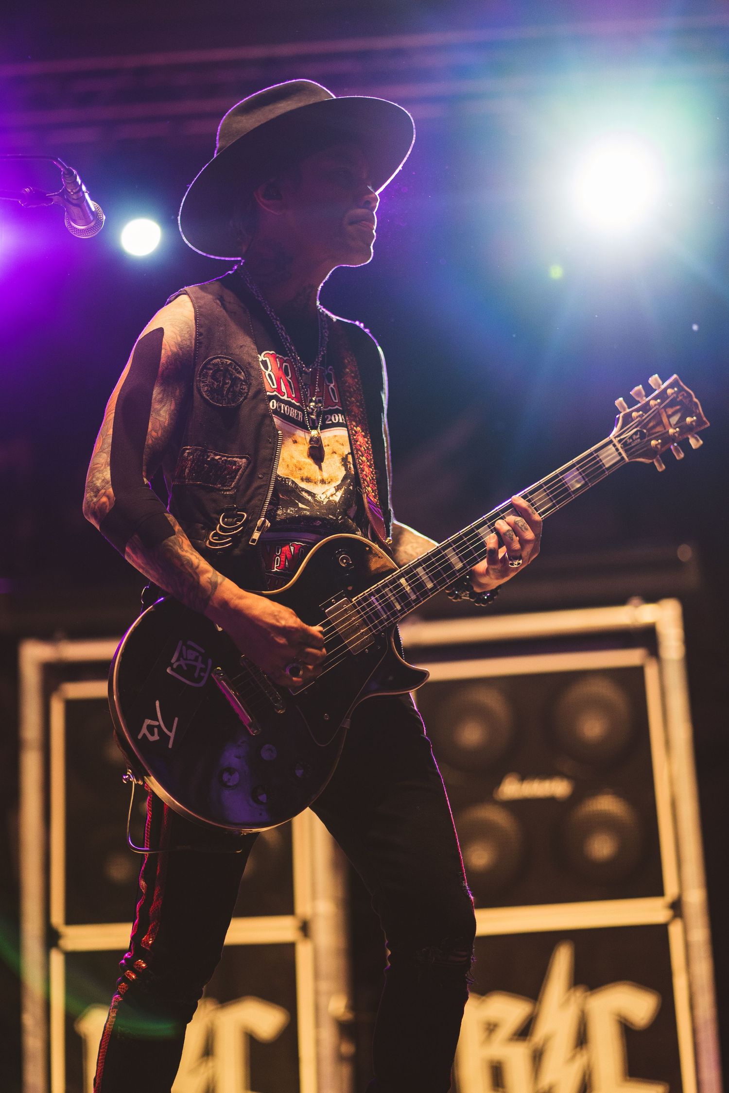 Musician in cowboy hat and vest plays electric guitar on concert stage with bright backlighting.