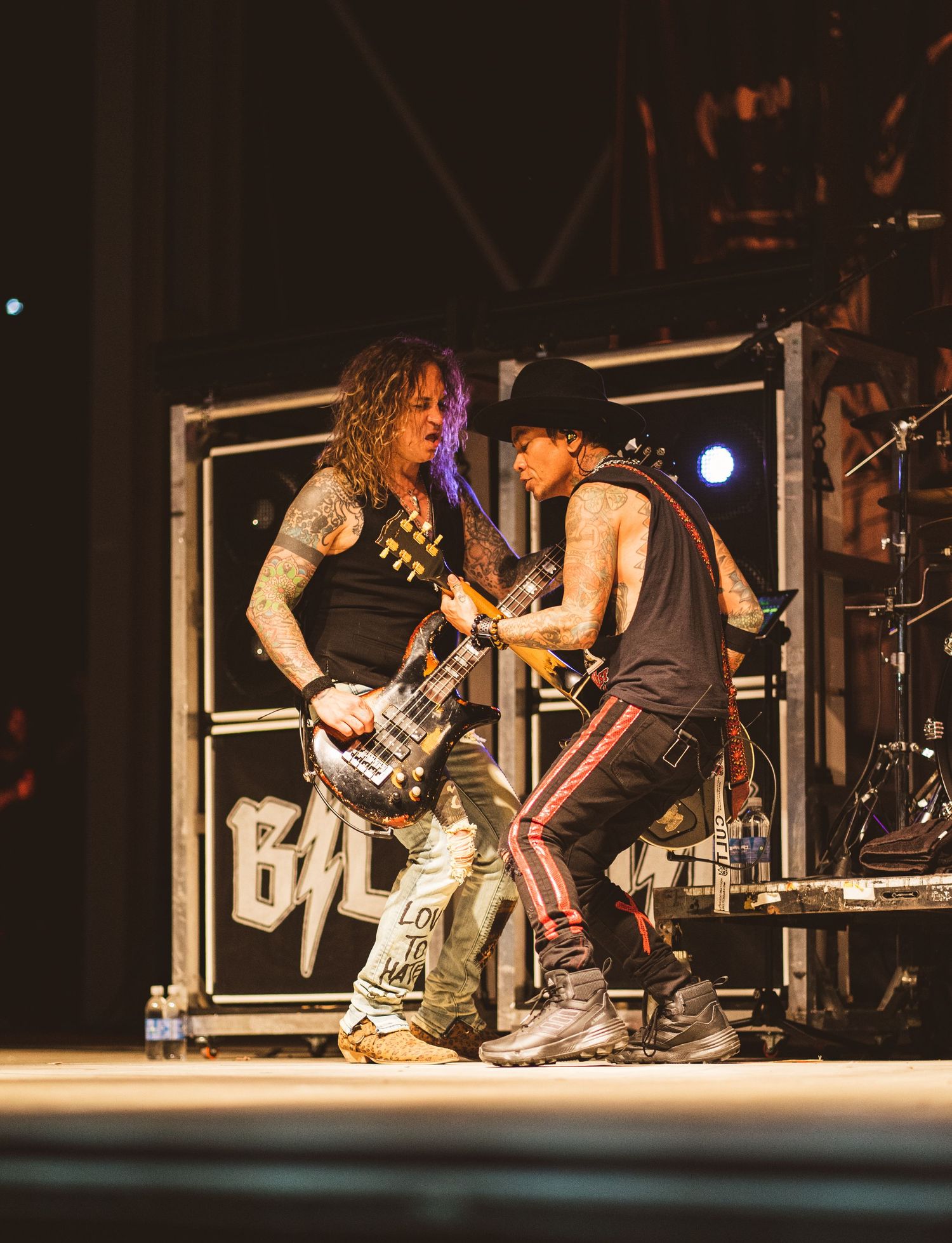 Two musicians perform energetically with guitars on stage in front of equipment stacks.