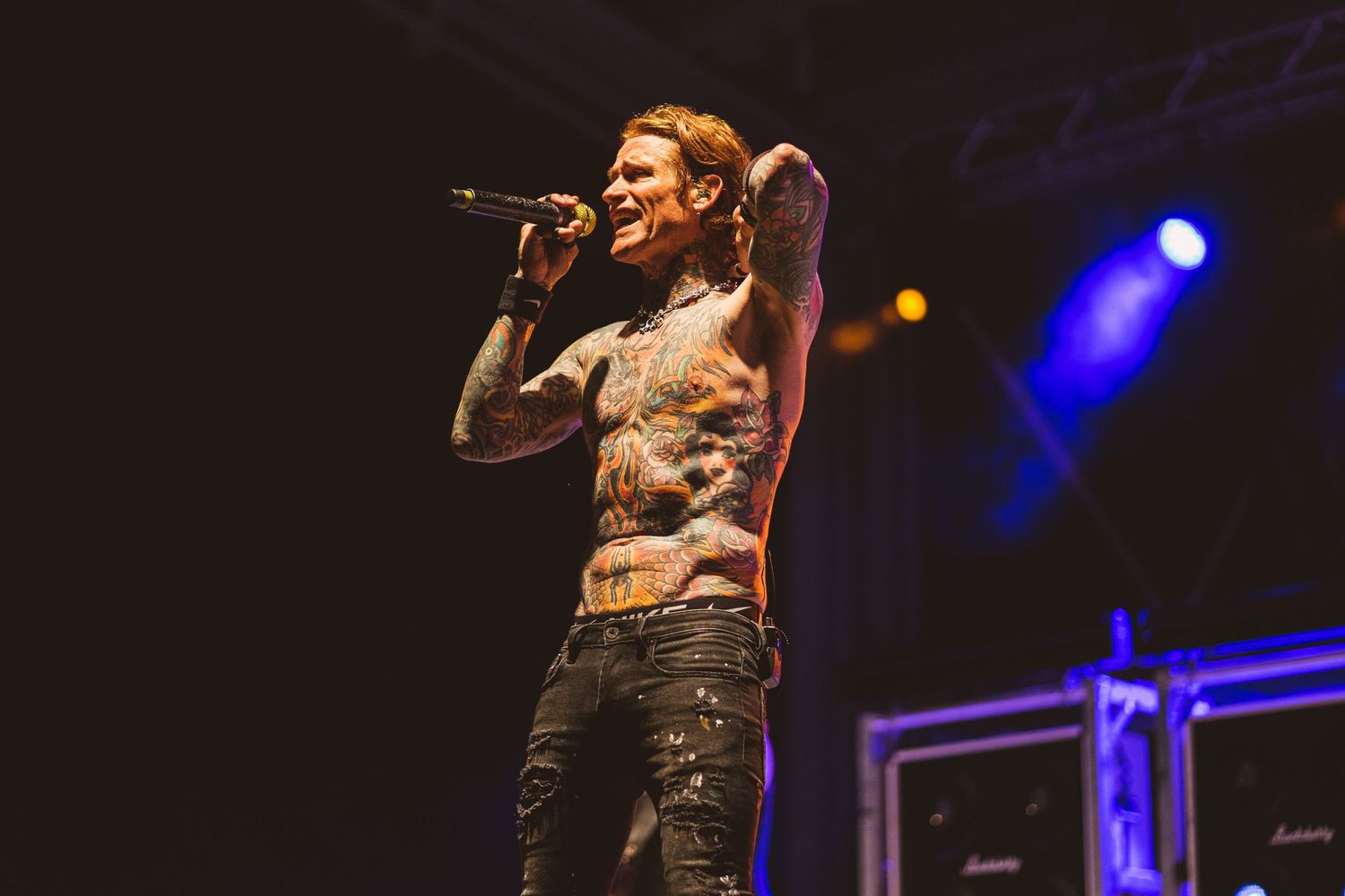 Shirtless performer with tattoos sings passionately on concert stage with blue lighting.