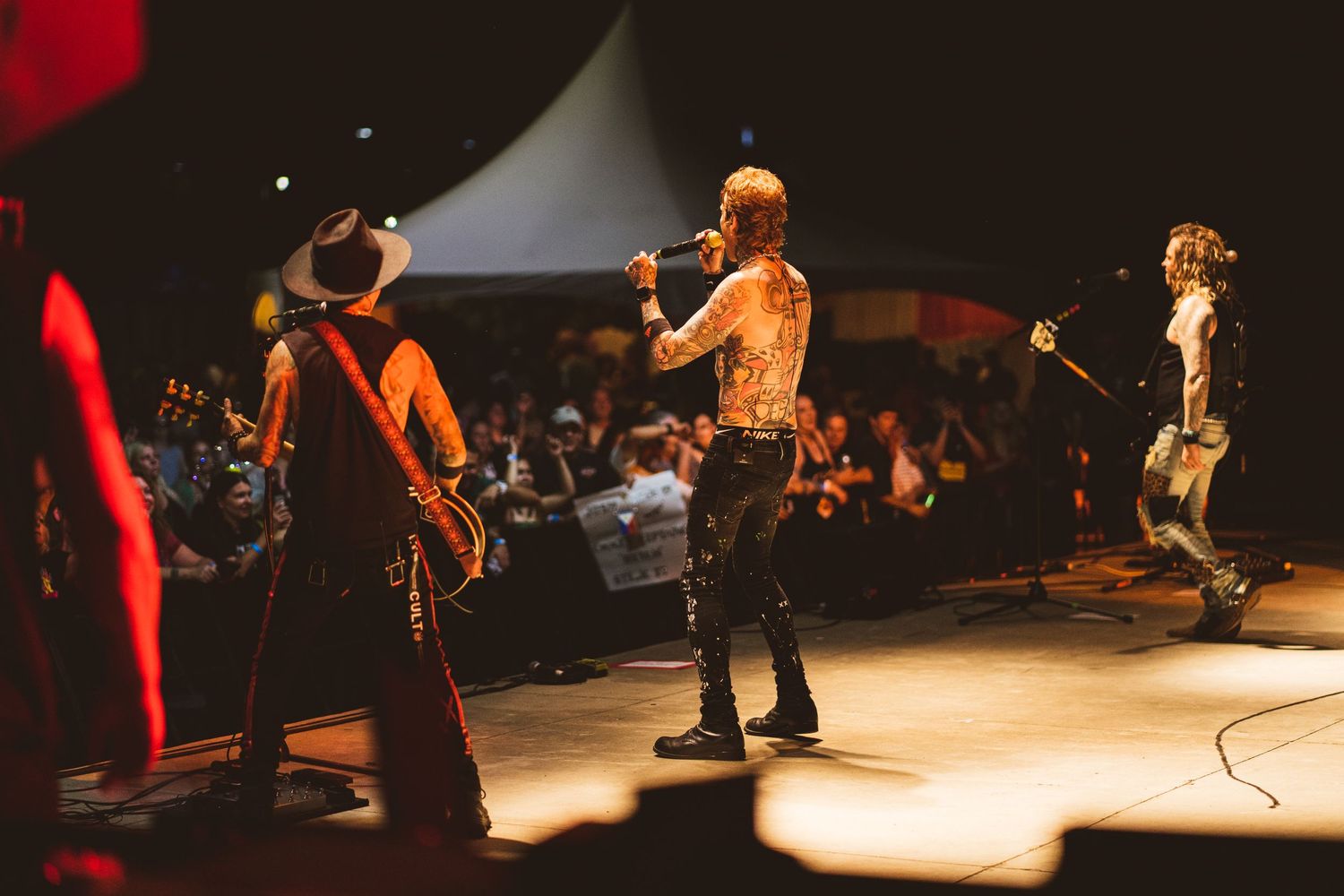 Band members perform on stage at outdoor concert venue with crowd visible.