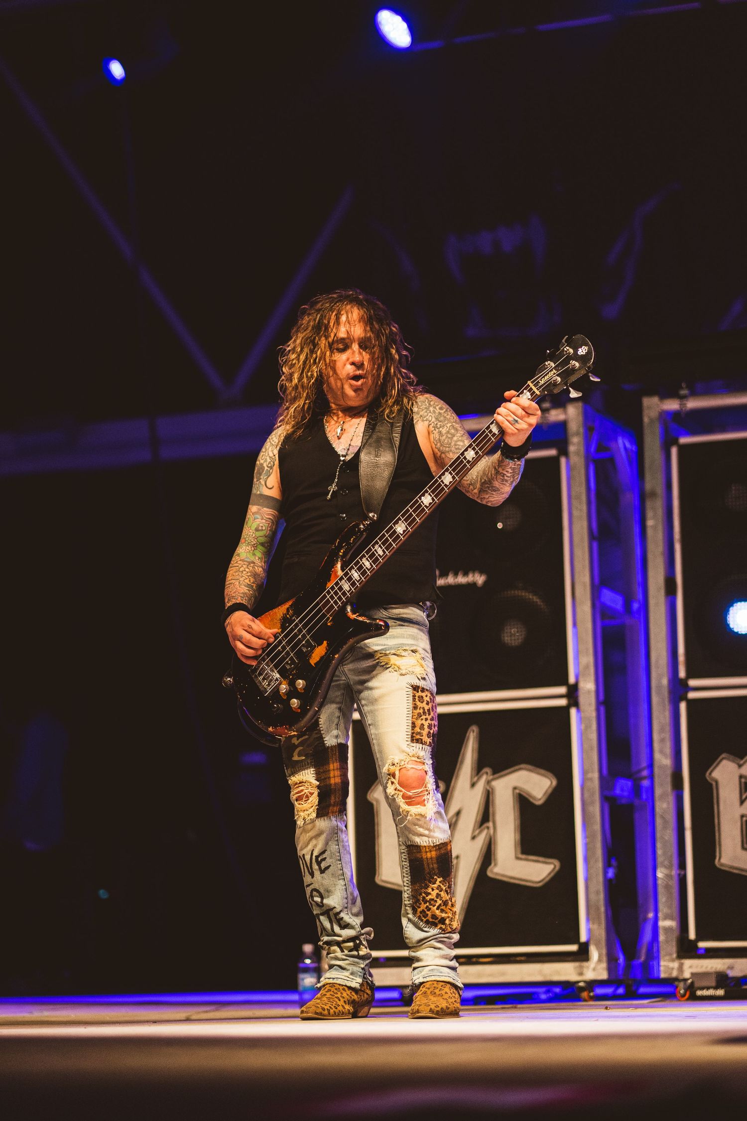 Bass guitarist with long hair and ripped jeans performs energetically on concert stage.