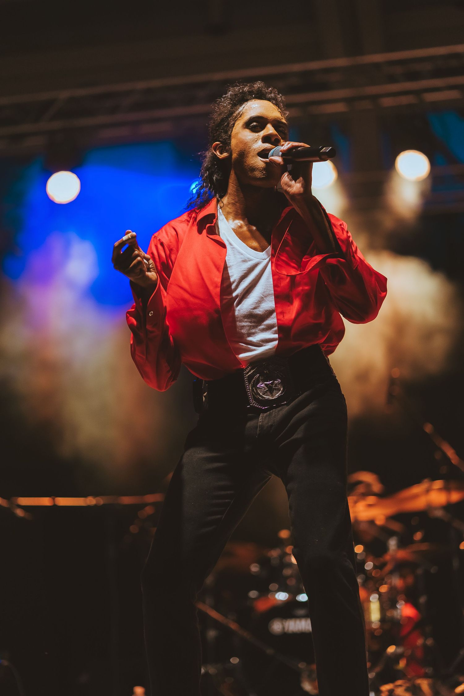 Performer in red jacket and black pants sings passionately on stage with blue lighting effects.