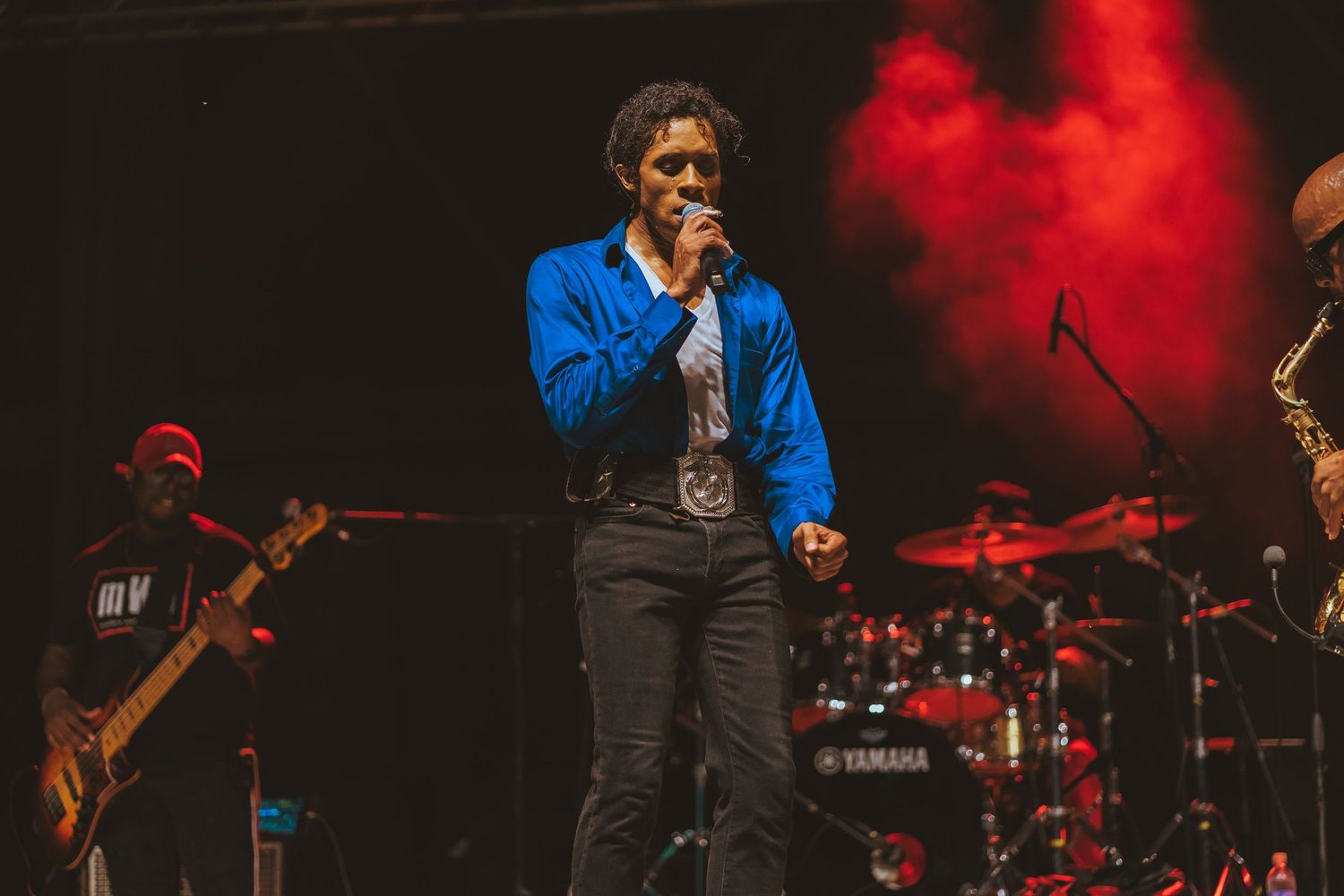 Stage performer in blue jacket sings with live band and dramatic red lighting in background.
