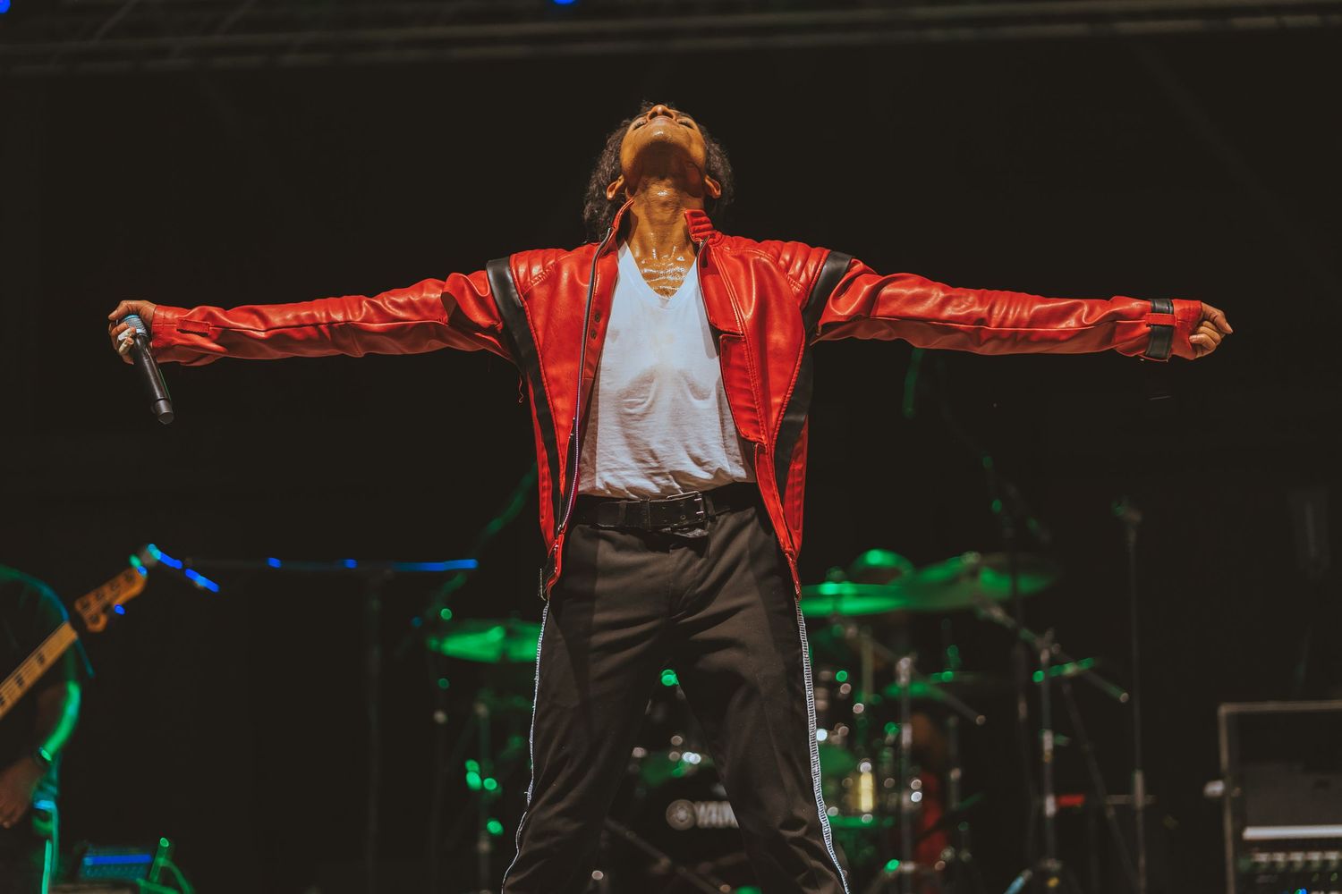 Performer in red leather jacket strikes dramatic pose with arms spread wide on concert stage.