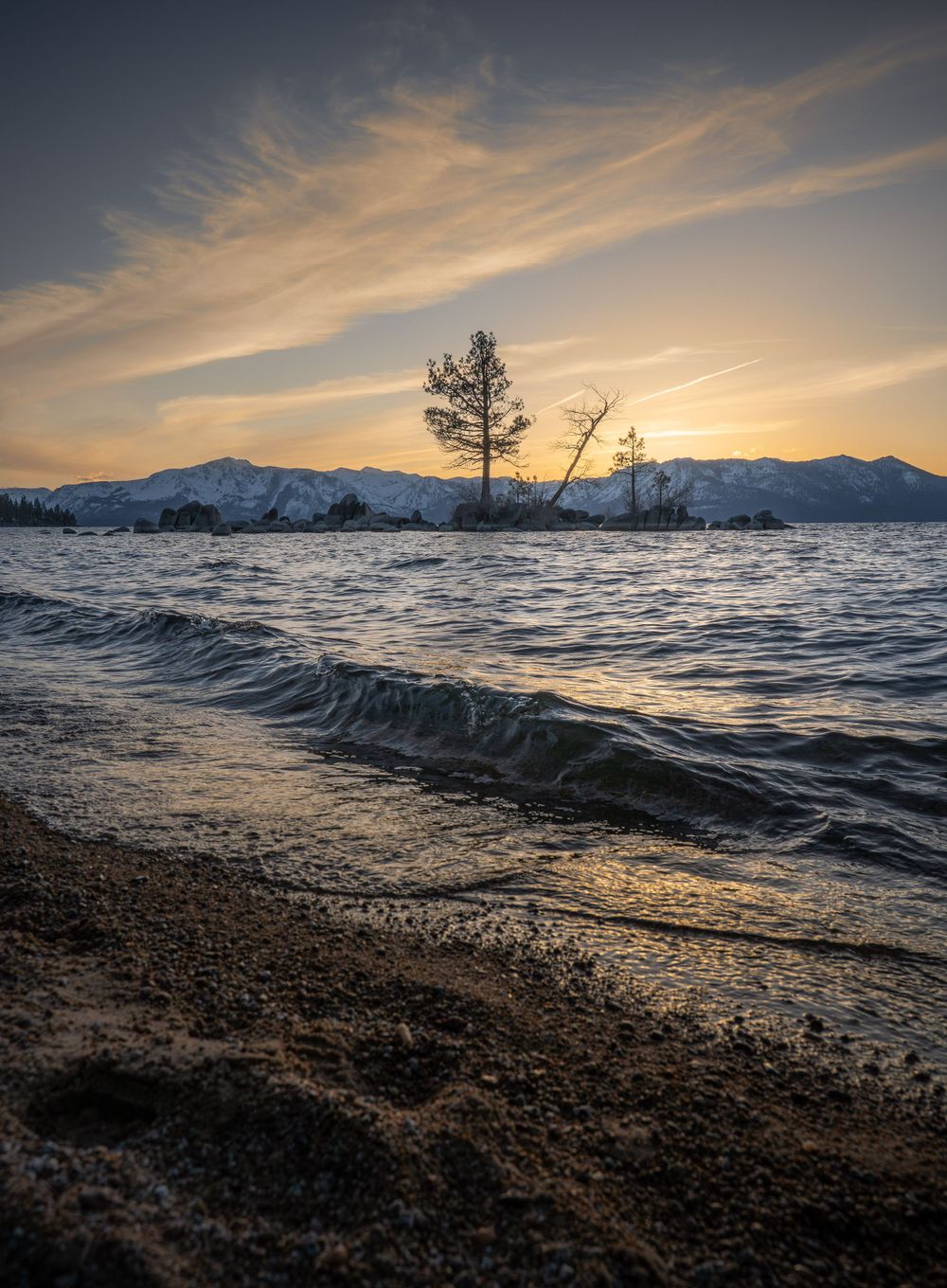 Dramatic sunset over Lake Tahoe with silhouetted tree and rolling waves crashing on sandy beach as mountains loom in background.