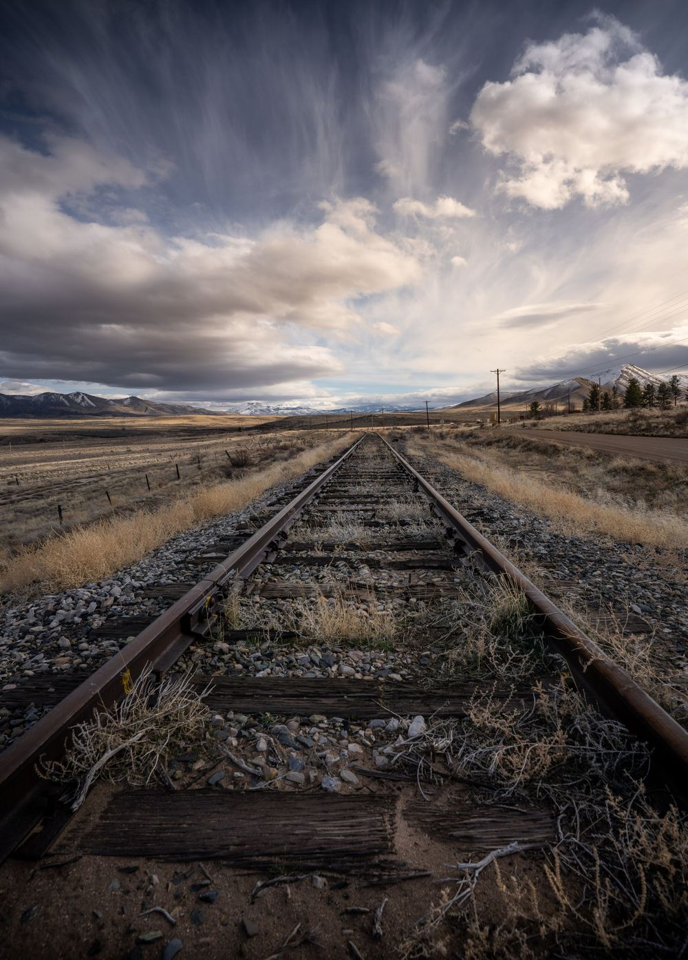 Old railroad tracks stretch into the distance under dramatic cloudy skies through a rural landscape with mountains on the horizon.