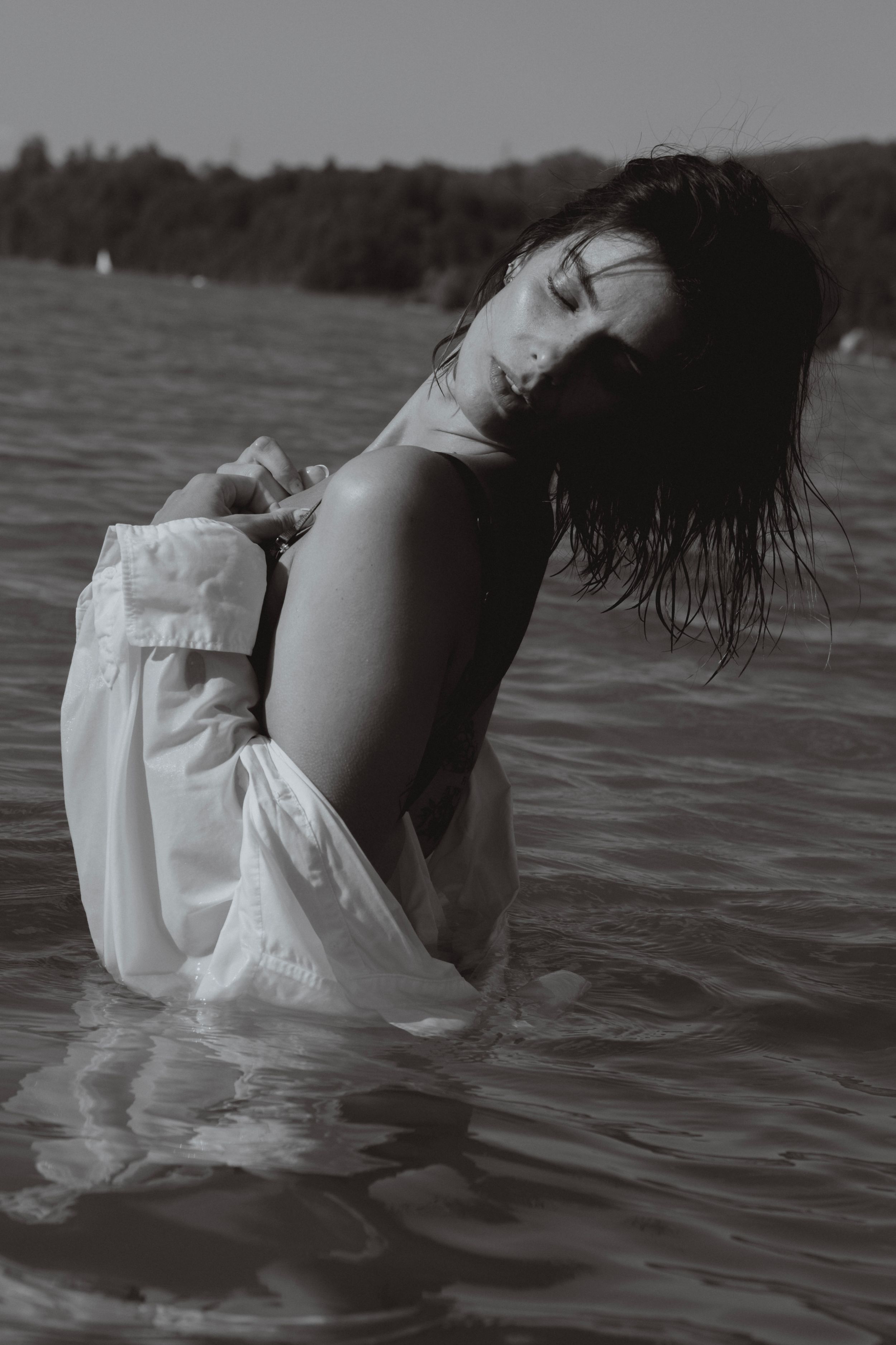 Black and white photo of a woman in a white dress standing in a lake with wet hair and eyes closed.