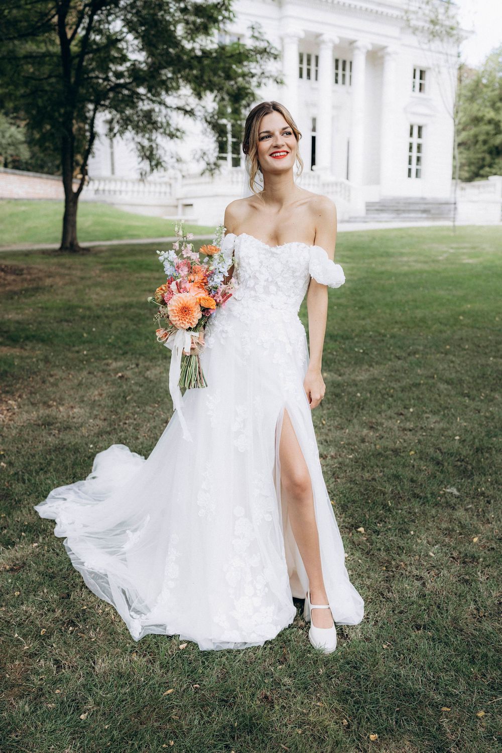 A bride in a flowing white lace gown poses gracefully on a grassy lawn with a peach and coral bouquet near a grand building.