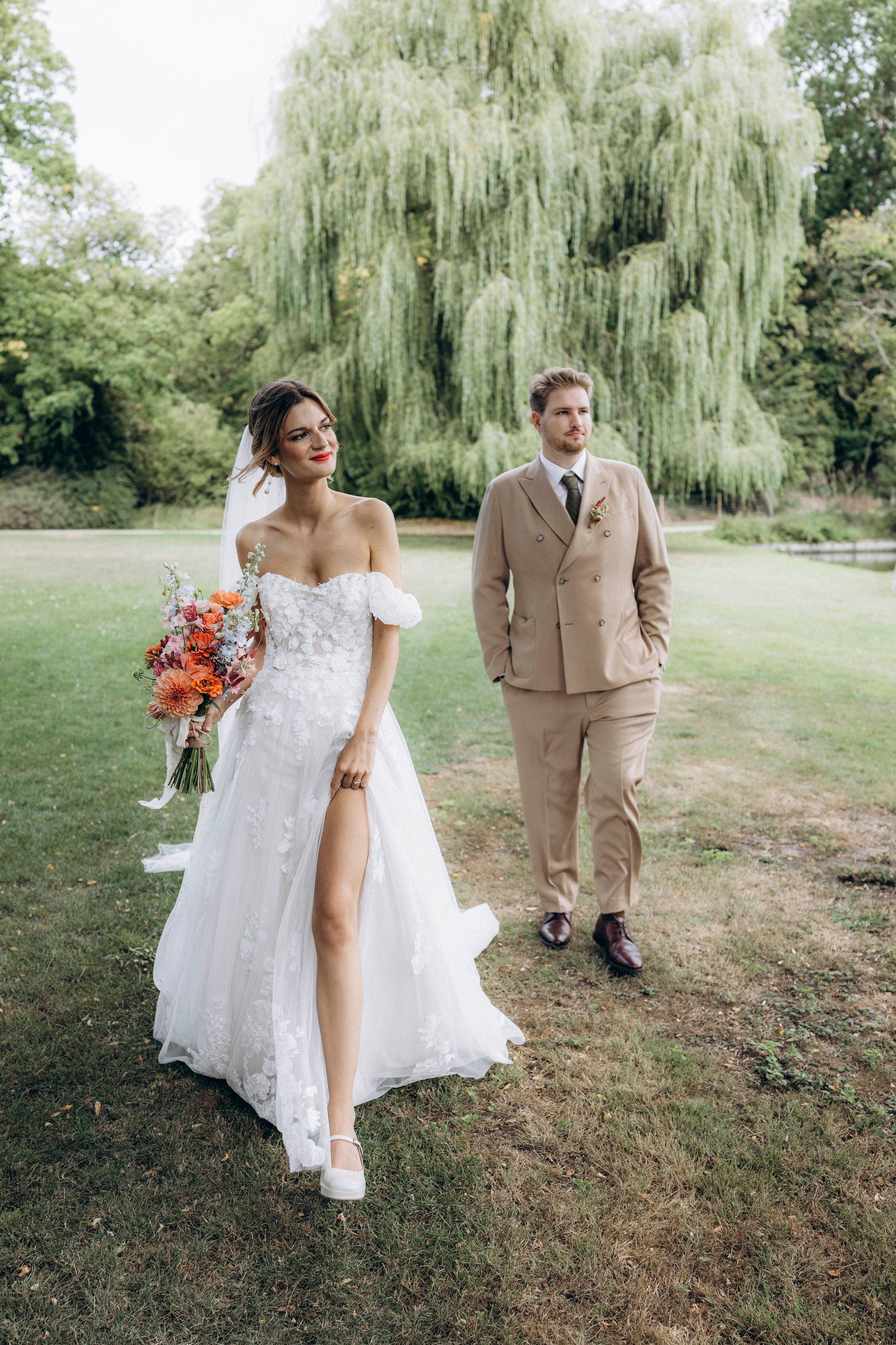 A couple in wedding attire poses together on a grassy lawn with a weeping willow tree in the background.