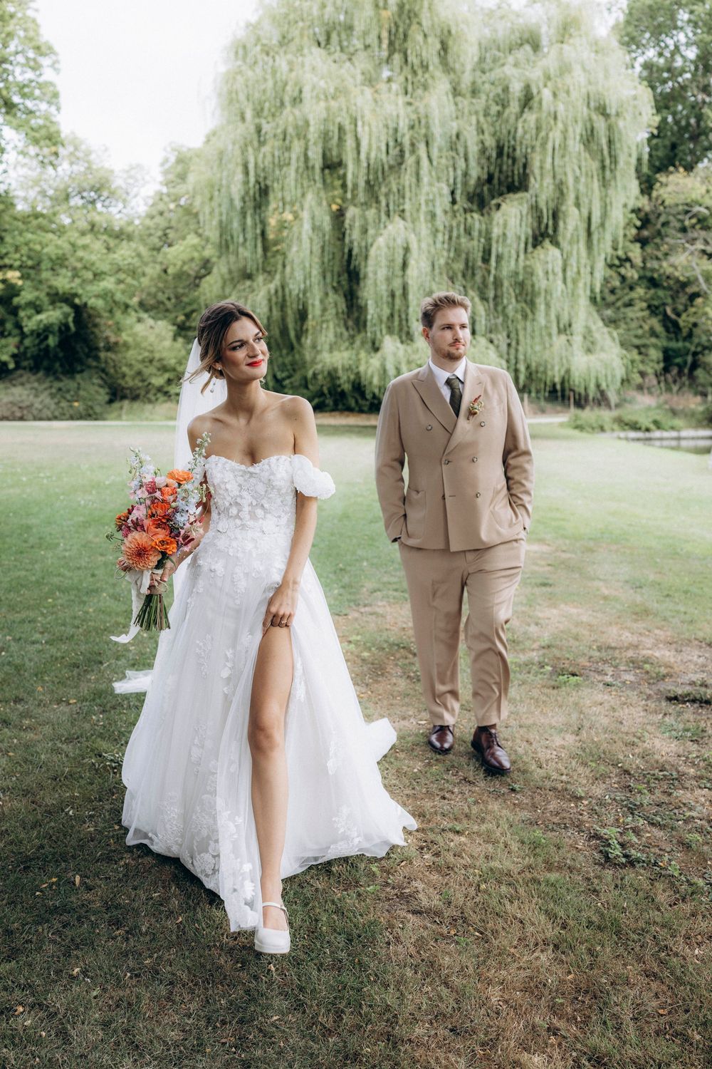 A couple in wedding attire poses together on a grassy lawn with a weeping willow tree in the background.