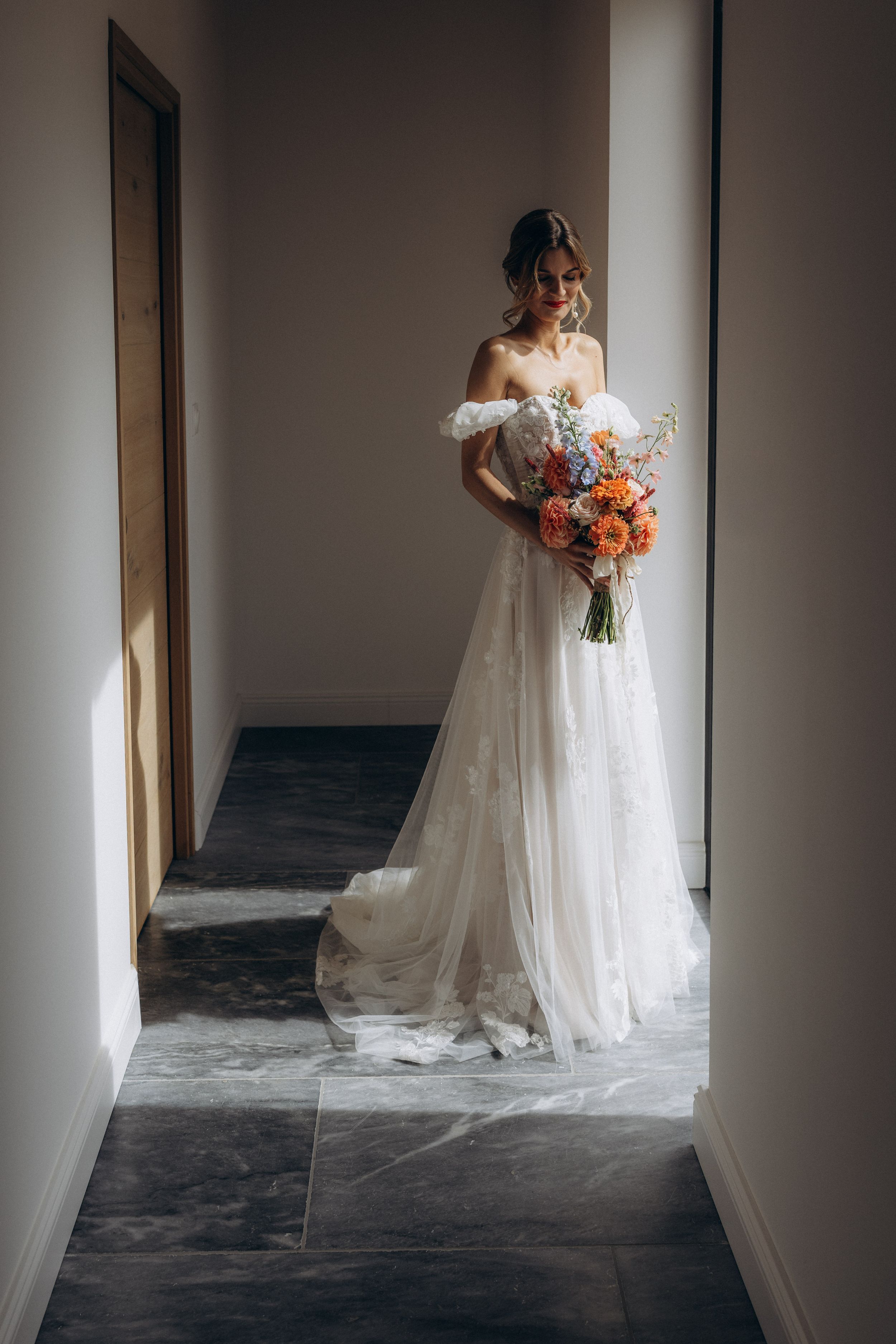 A bride in a flowing white wedding dress holds a vibrant coral and peach bouquet while standing in a minimalist hallway.