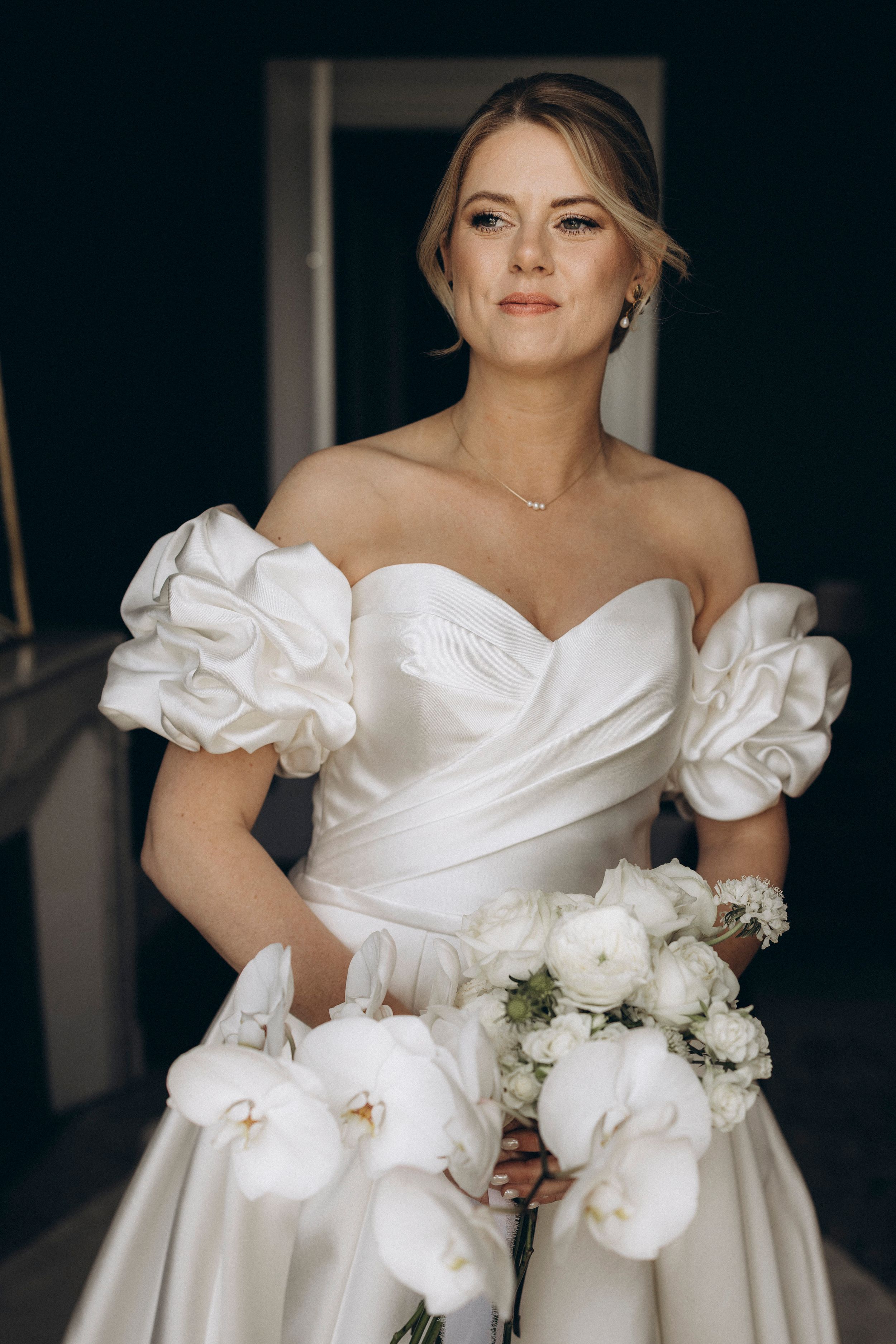 Elegant bride in off-shoulder white wedding gown with puff sleeves and white floral bouquet in moody lighting.