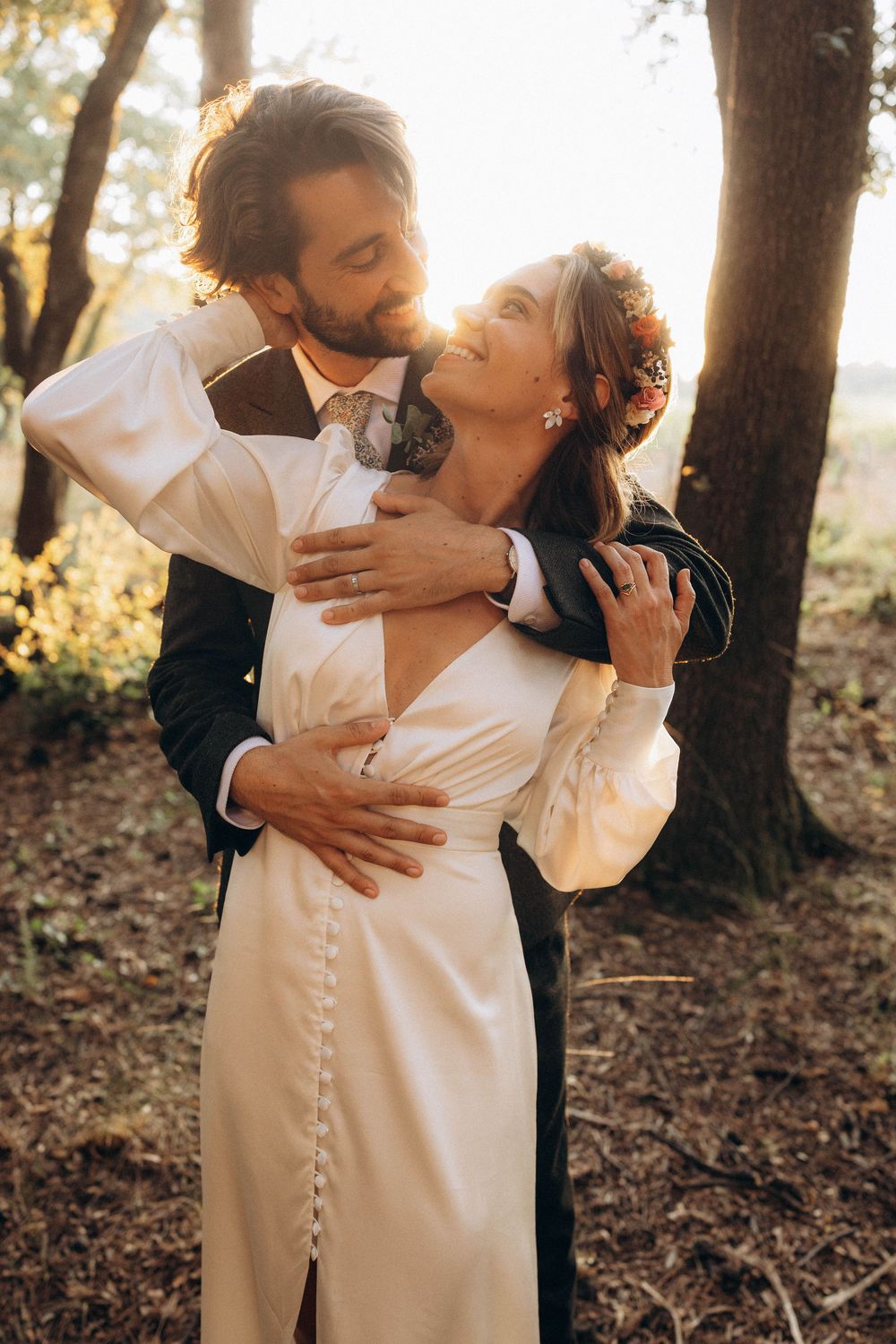 A romantic couple embraces in a sunlit forest setting wearing elegant white wedding attire.