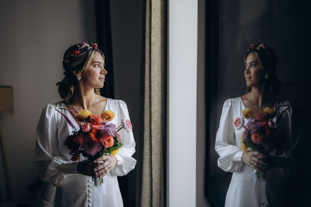 A person in a white dress holds a colorful bouquet of flowers while posing near a window in moody lighting.