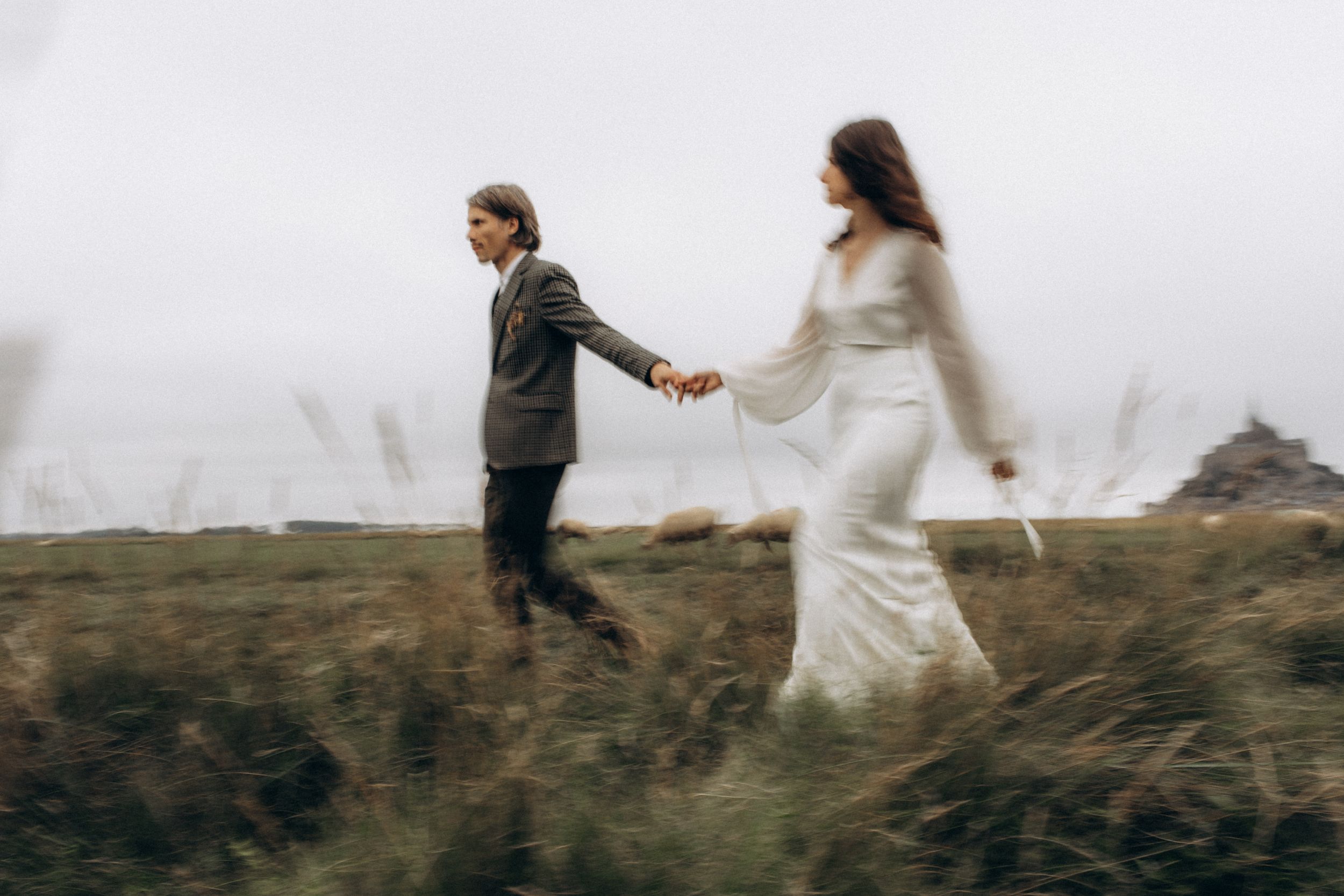 A couple runs hand in hand through a misty field, with the bride wearing a flowing white dress and groom in dark attire.