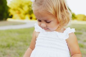 Young child in white dress looking down while standing outdoors in natural lighting.