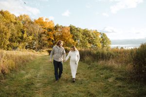 Couple walking hand in hand through autumn field with golden trees in background.