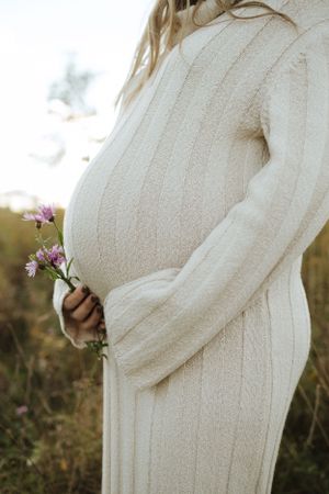 Pregnant woman in white sweater dress holding purple wildflowers against sunset backdrop.
