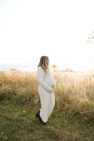 Expectant mother in white dress standing in golden field during sunset maternity photoshoot.