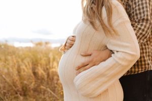 Close-up of pregnant belly in white sweater dress with partner's hands embracing from behind.