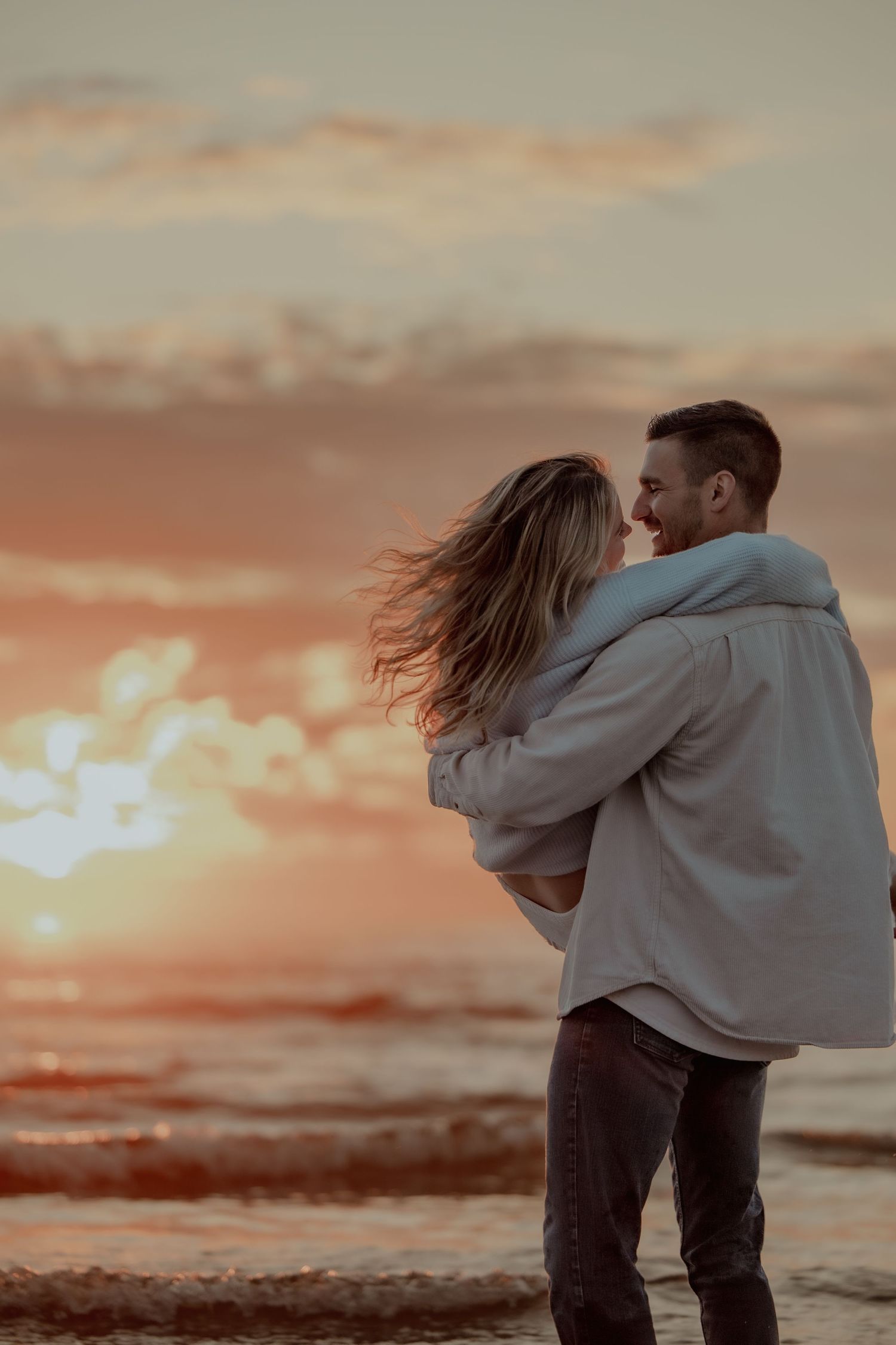 A couple embraces on a beach at sunset with dramatic orange sky and windswept hair creating a romantic silhouette.
