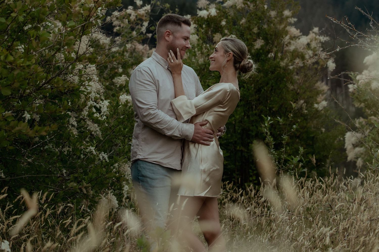 A couple embraces romantically in a sunlit meadow surrounded by tall grass and wildflowers during a golden hour photoshoot.