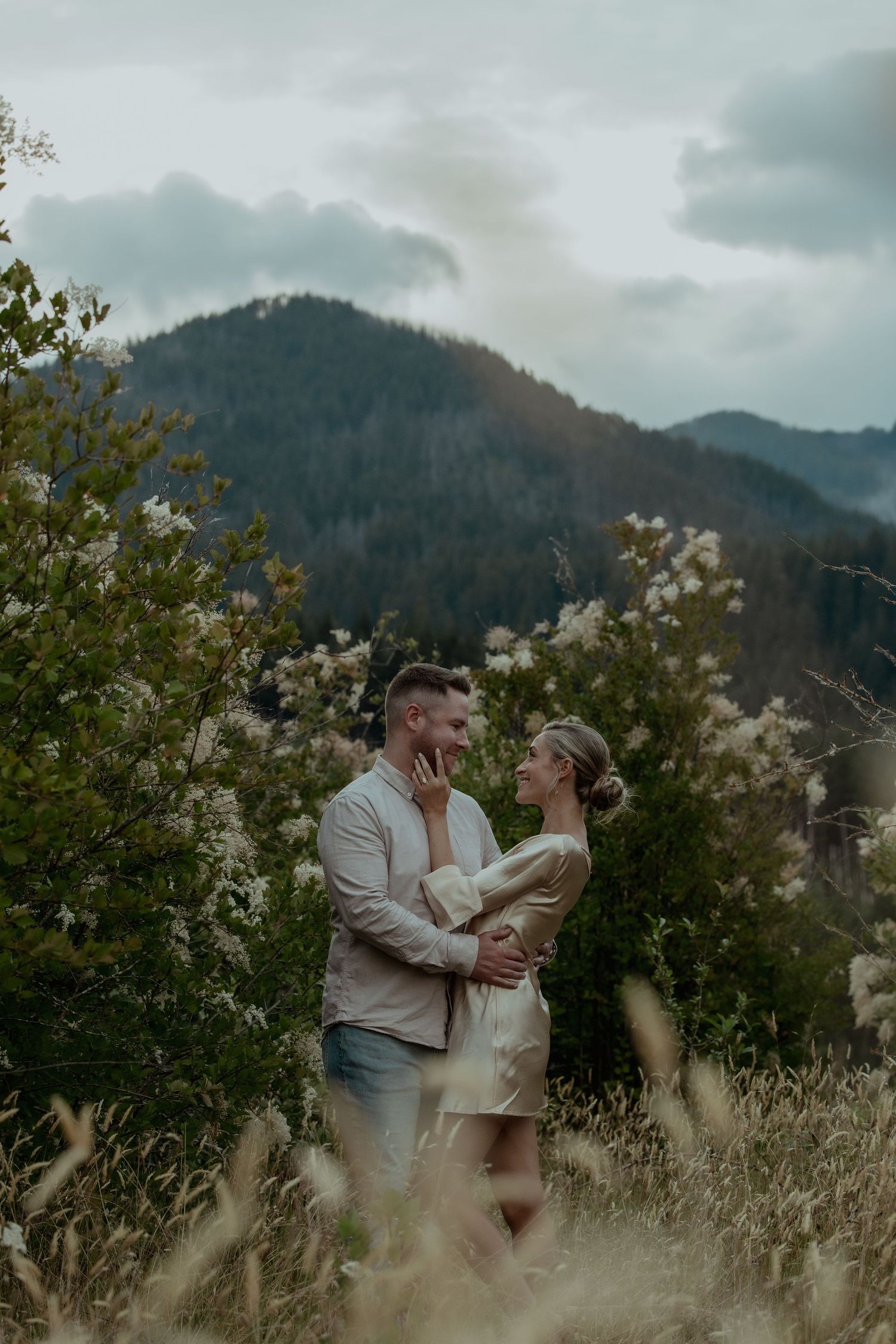 A romantic couple embraces in a field with misty mountains and moody clouds in the background.