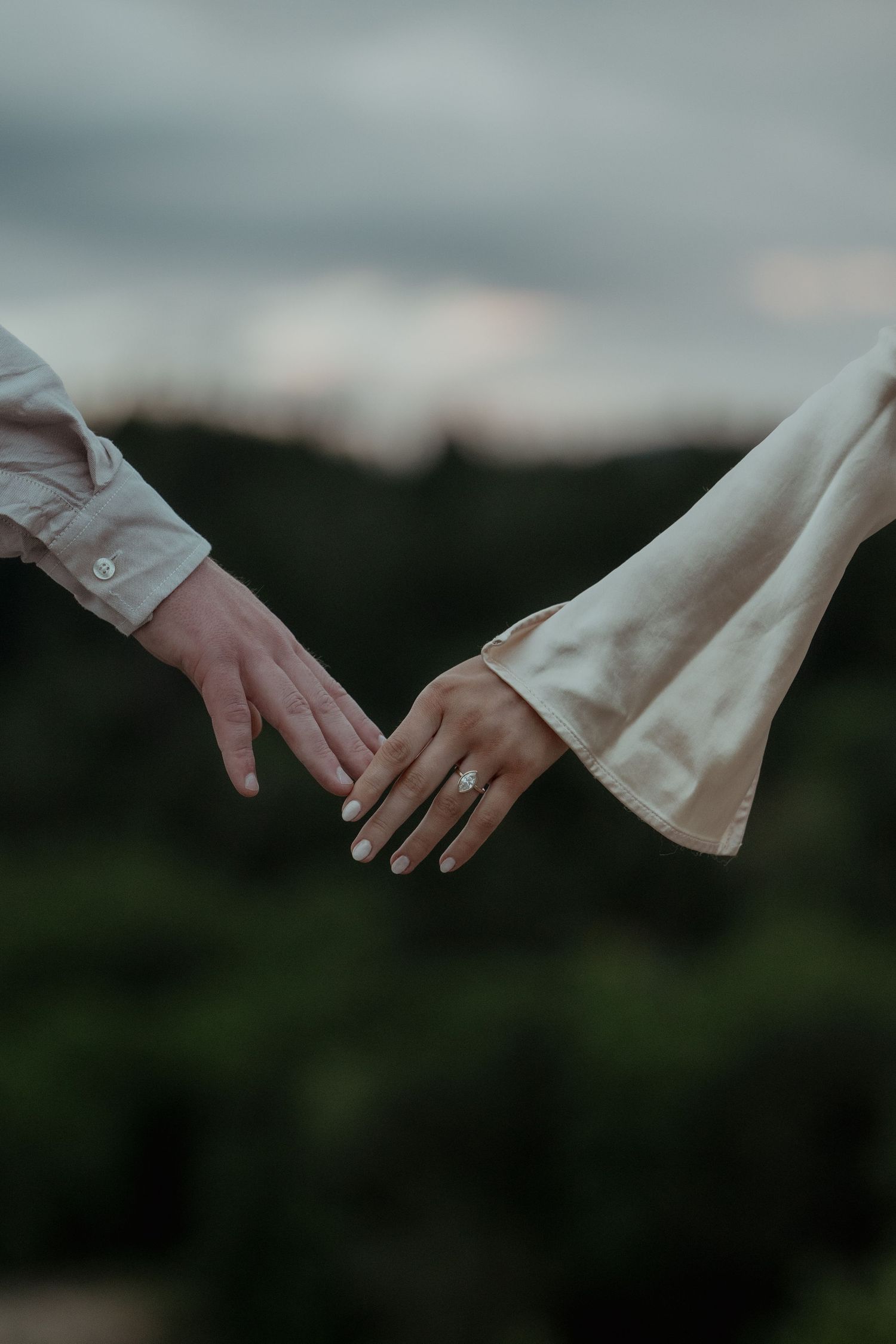 Two hands reaching towards each other with one wearing an engagement ring against a moody outdoor backdrop at dusk.