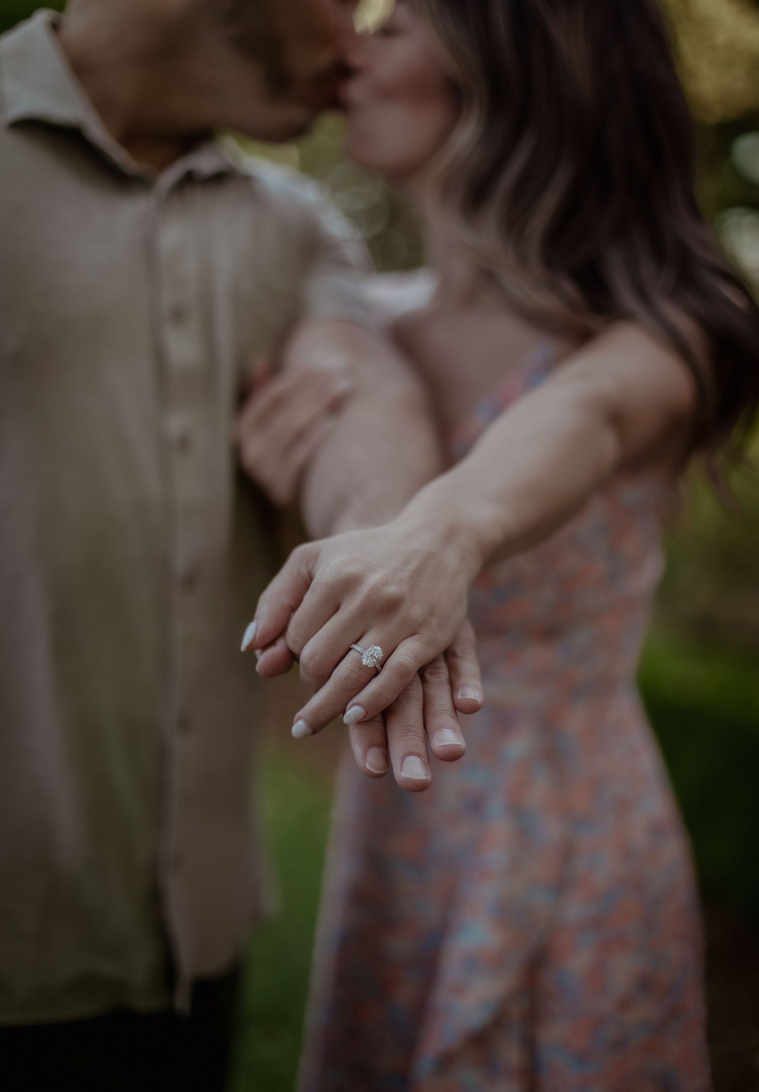 A person wearing a polka dot dress shows off an engagement ring in a romantic outdoor setting.