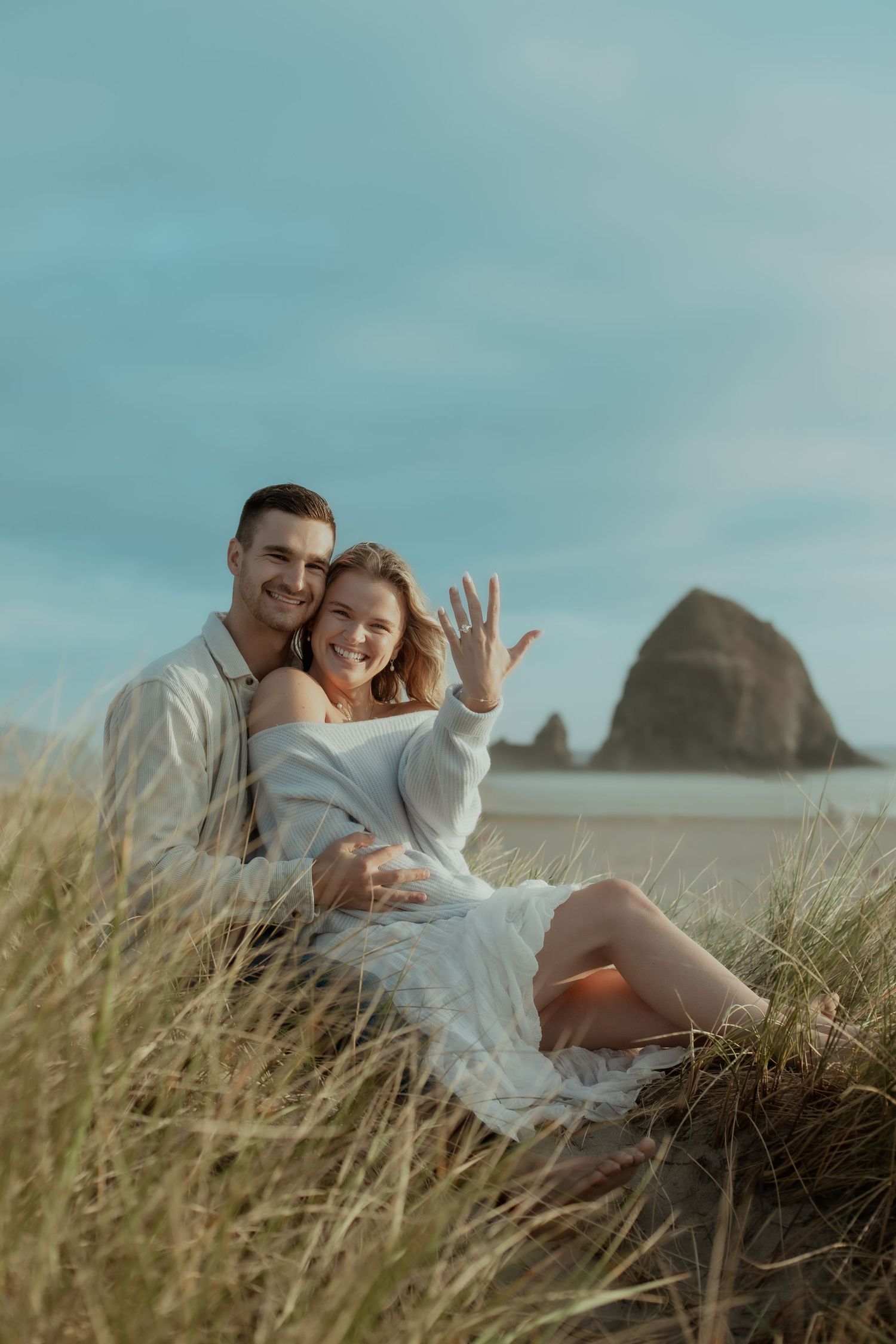 A couple embraces on a grassy hillside overlooking a misty beach landscape with dramatic coastal cliffs in the background.