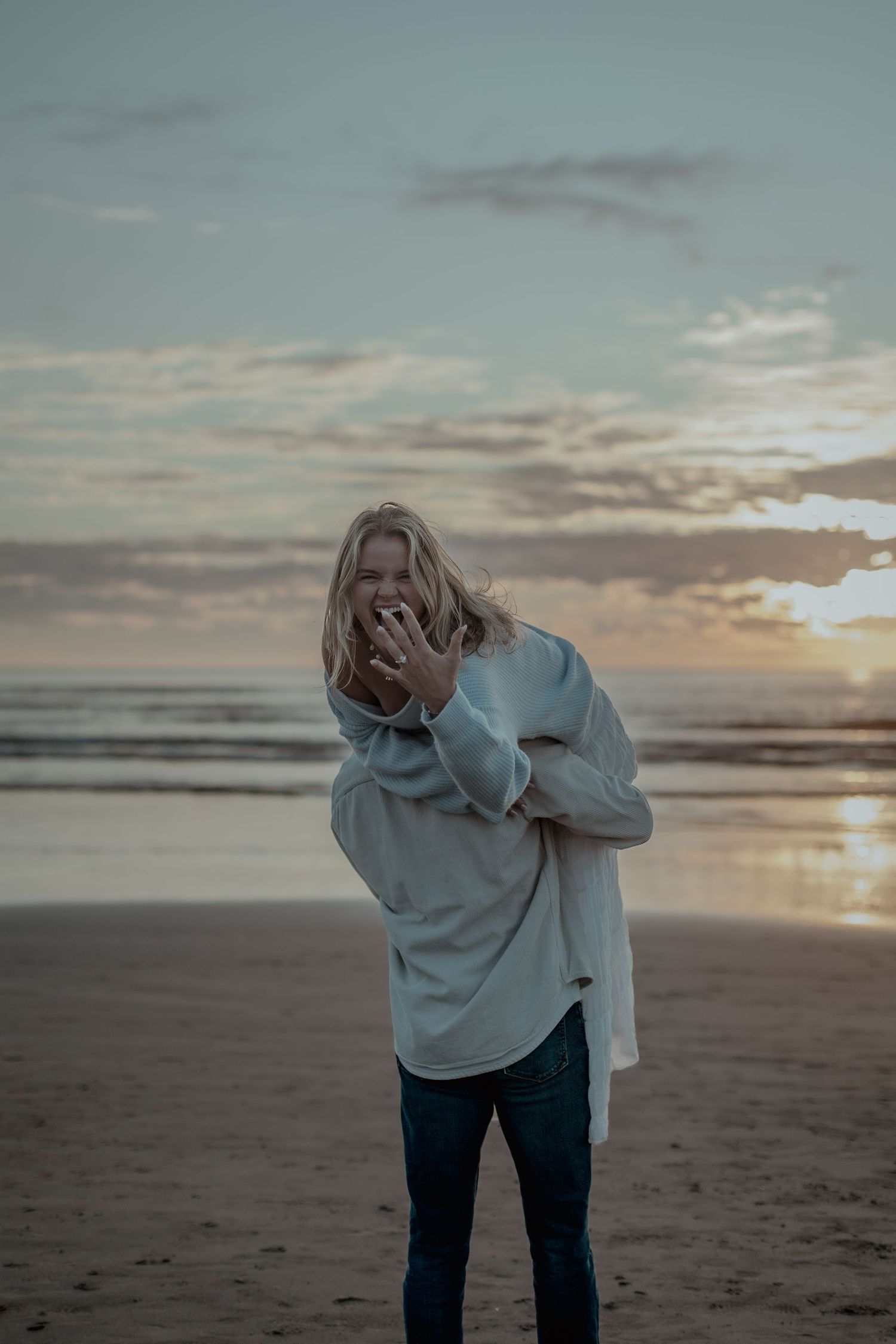 A person in a light colored sweater stands on a misty beach at sunset with emotional body language.